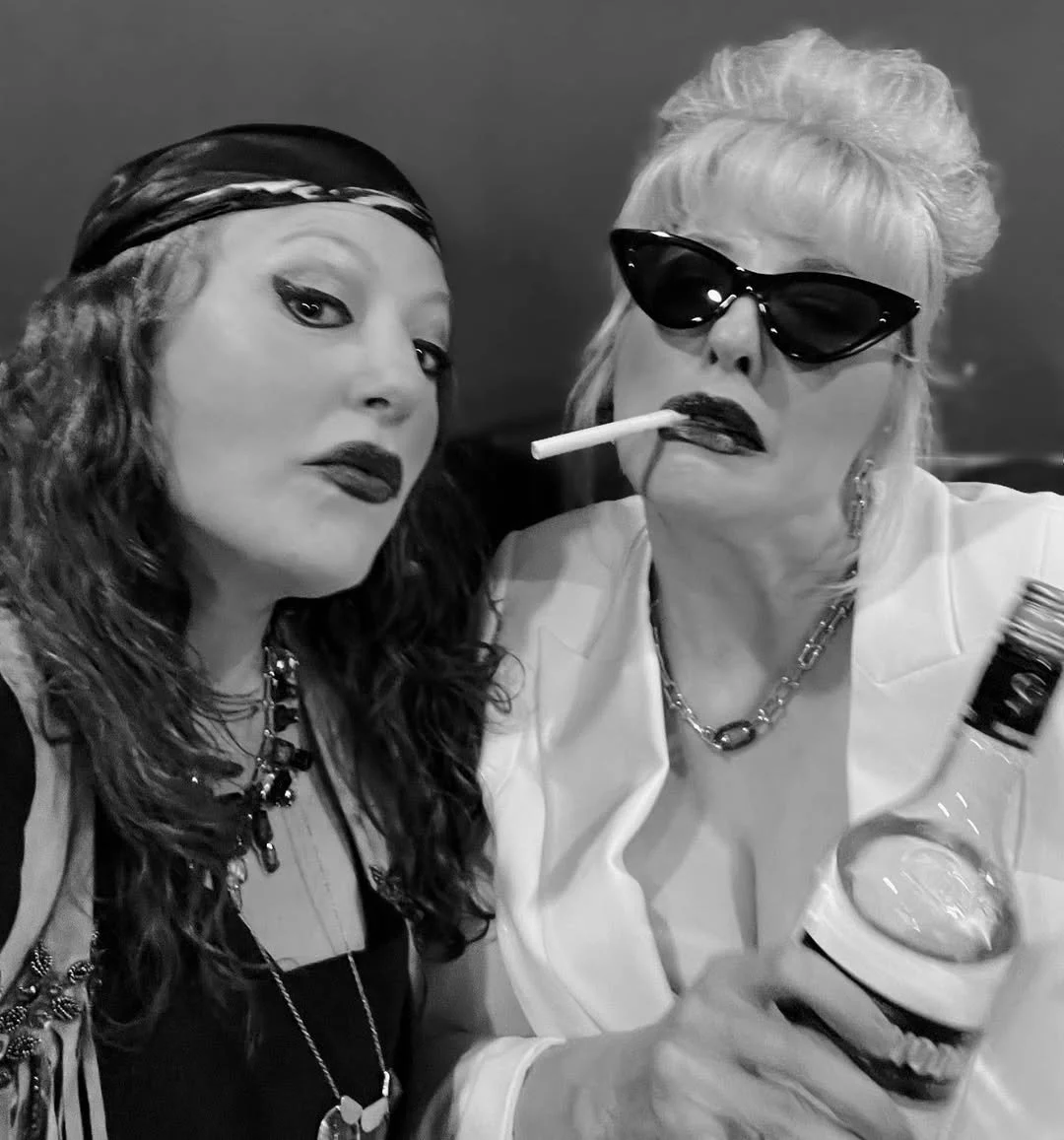 Two women with alternative fashion styles, one with dark lipstick and a headband, the other wearing sunglasses, smoking a cigarette in a bottle, and dressed in white.