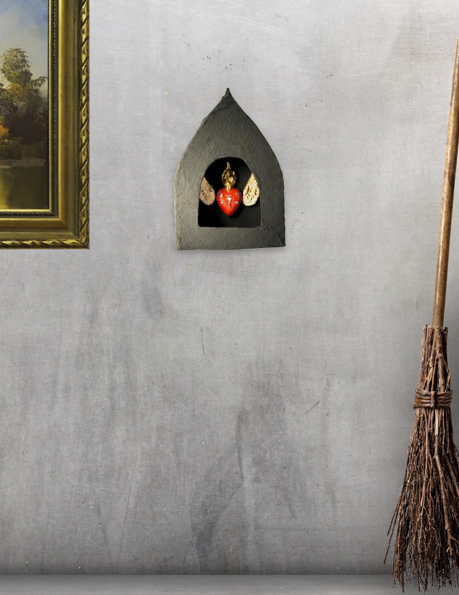 A small black house-shaped shelf with a decorative angel holding a red heart with white dots and gold wings inside; part of a painting in a gold frame is visible on the left, and a bundle of sticks tied with wire is on the right side.