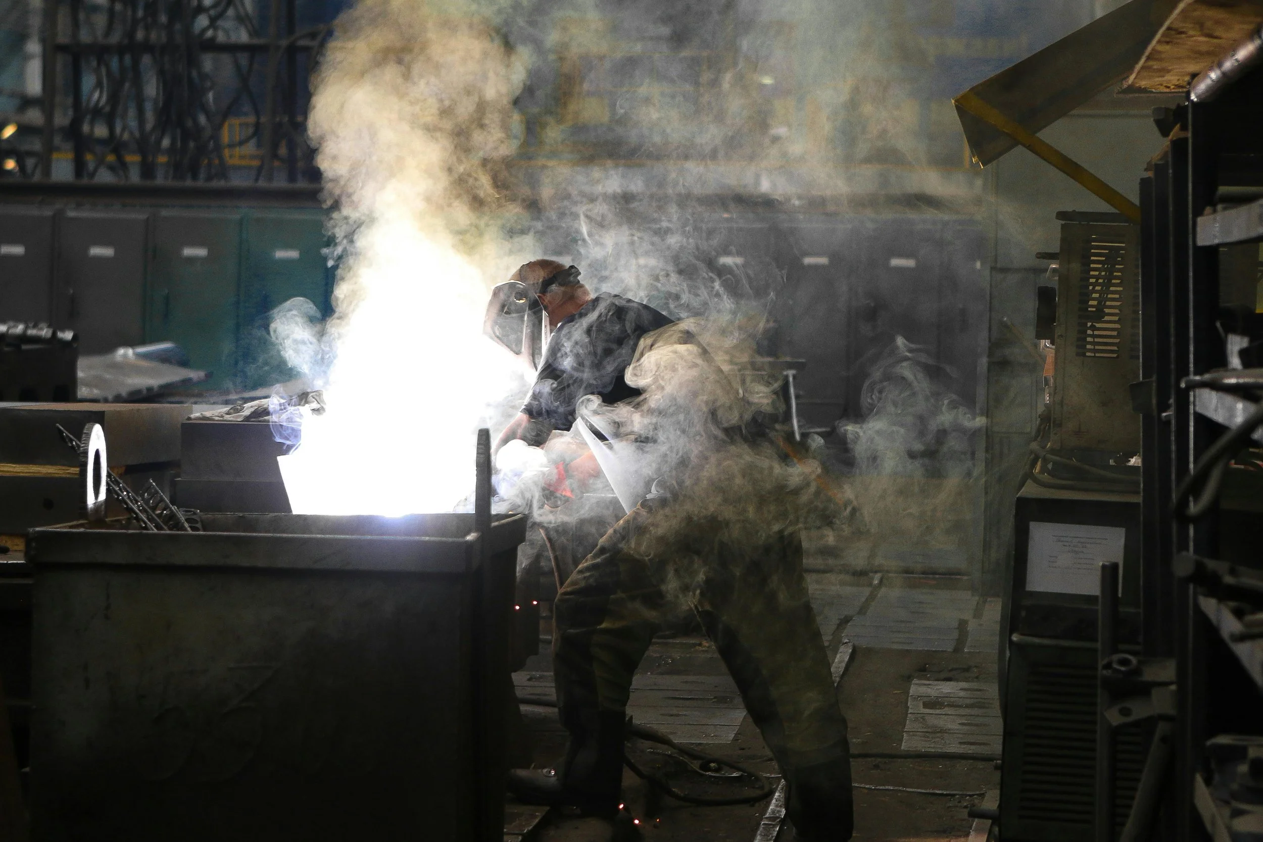 A person welding in a workshop surrounded by sparks and smoke, wearing protective gear including a welding helmet and gloves.