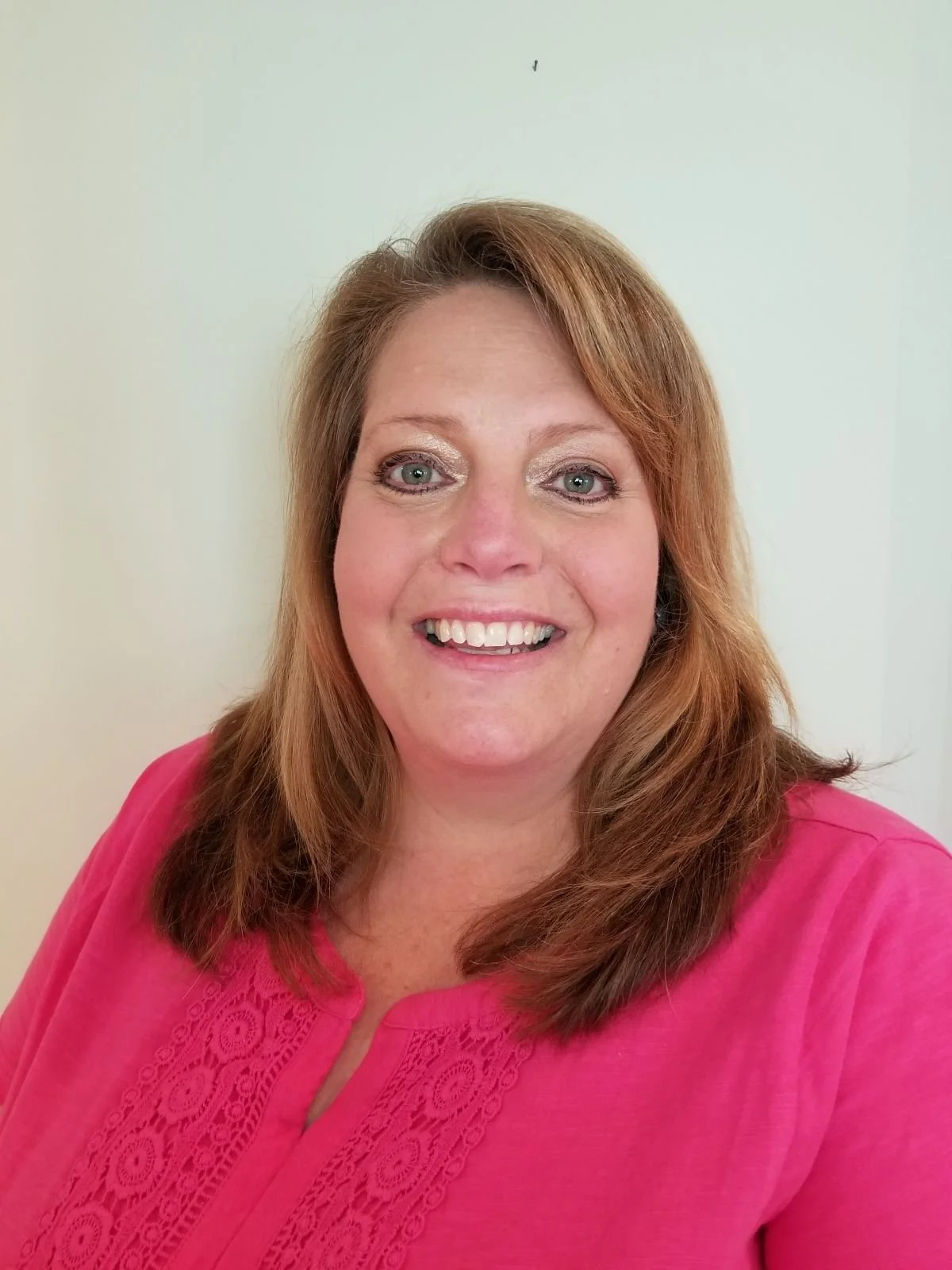 Portrait of a woman with red hair smiling, wearing a pink blouse, standing against a plain light-colored background.