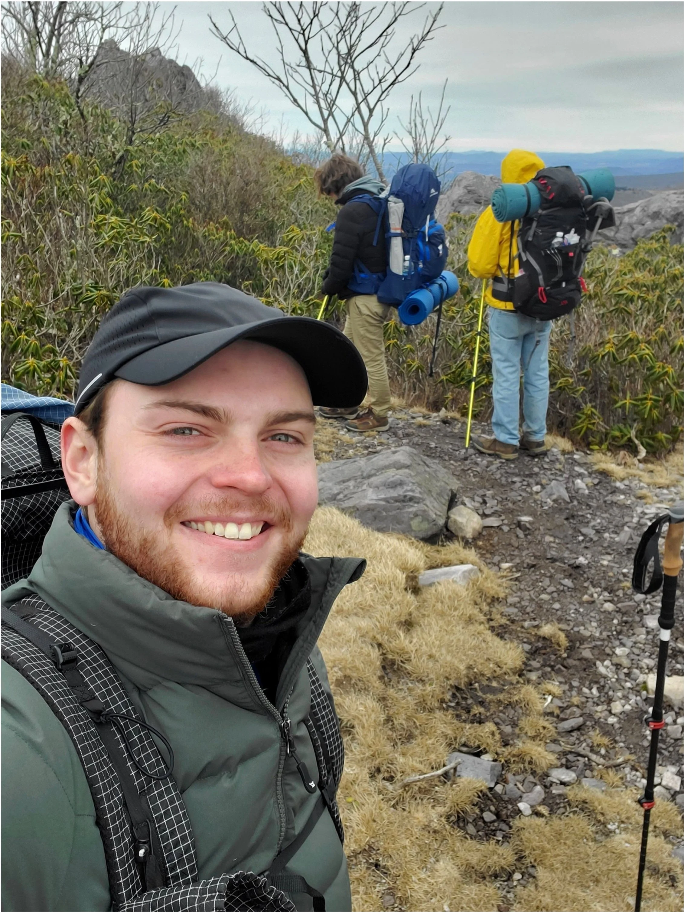 Three hikers on a mountain trail, with a man smiling taking a selfie in the foreground, and two people with backpacks and trekking poles looking at the view in the background.