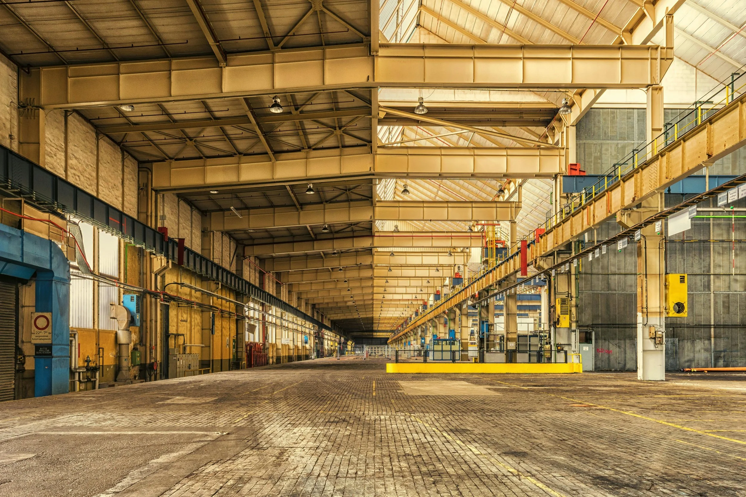 Empty industrial warehouse interior with metal beams, overhead cranes, and a wide brick floor.