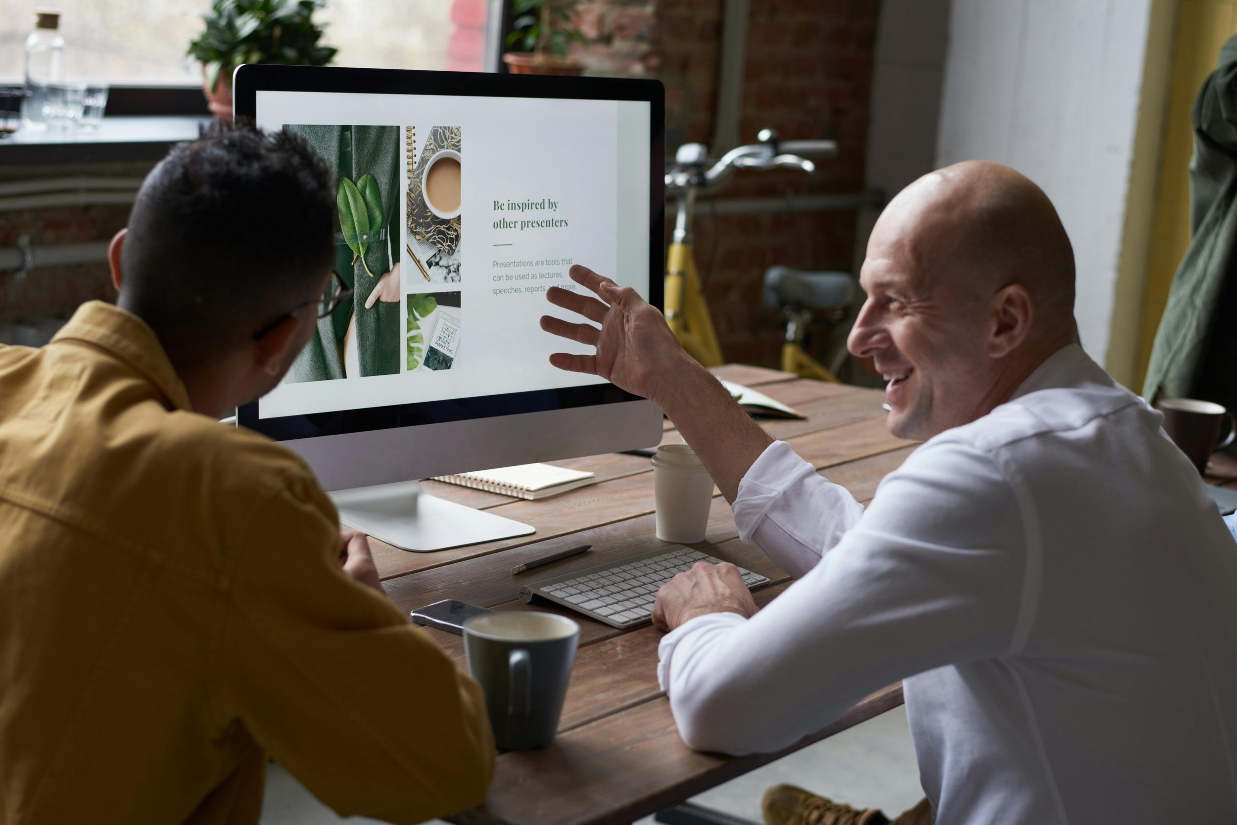 Two men sitting at a wooden table, looking at a computer screen with a presentation slide. One is pointing at the screen and smiling, the other is listening. The setting appears to be an office or coworking space with a brick wall and a bicycle in the background.