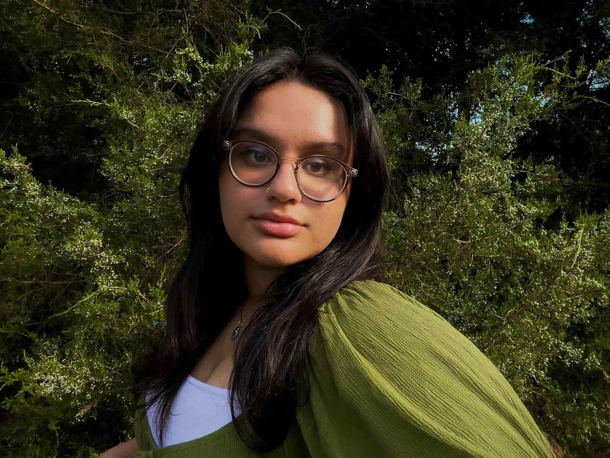 Mina Hakimpour a creative director in Washington DC has with long dark hair and glasses stands outdoors in front of green bushes, wearing a green top and white shirt.