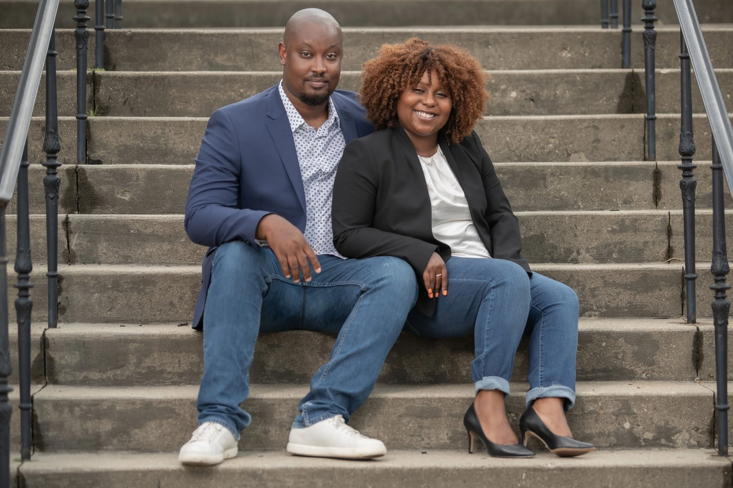 A man and woman sitting on outdoor concrete stairs, smiling at the camera. The man wears a blue blazer, white patterned shirt, blue jeans, and white sneakers. The woman wears a black blazer, white blouse, blue jeans, and black high heels.