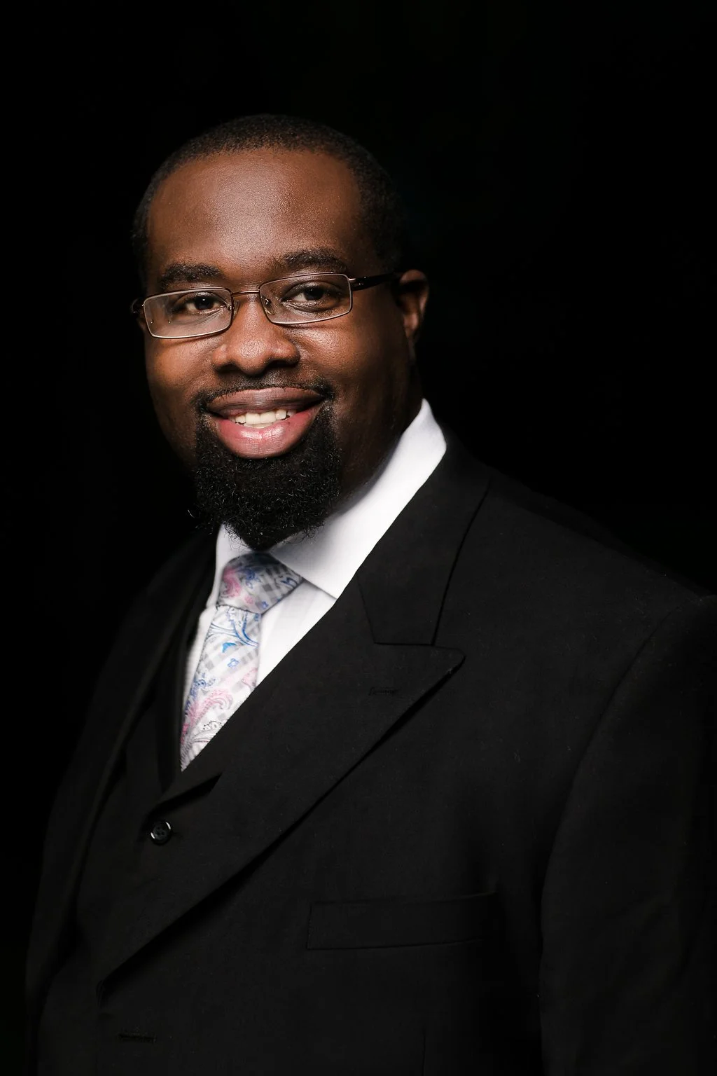 Portrait of a smiling Black man in a black suit, white shirt, and paisley tie, wearing glasses against a black background.