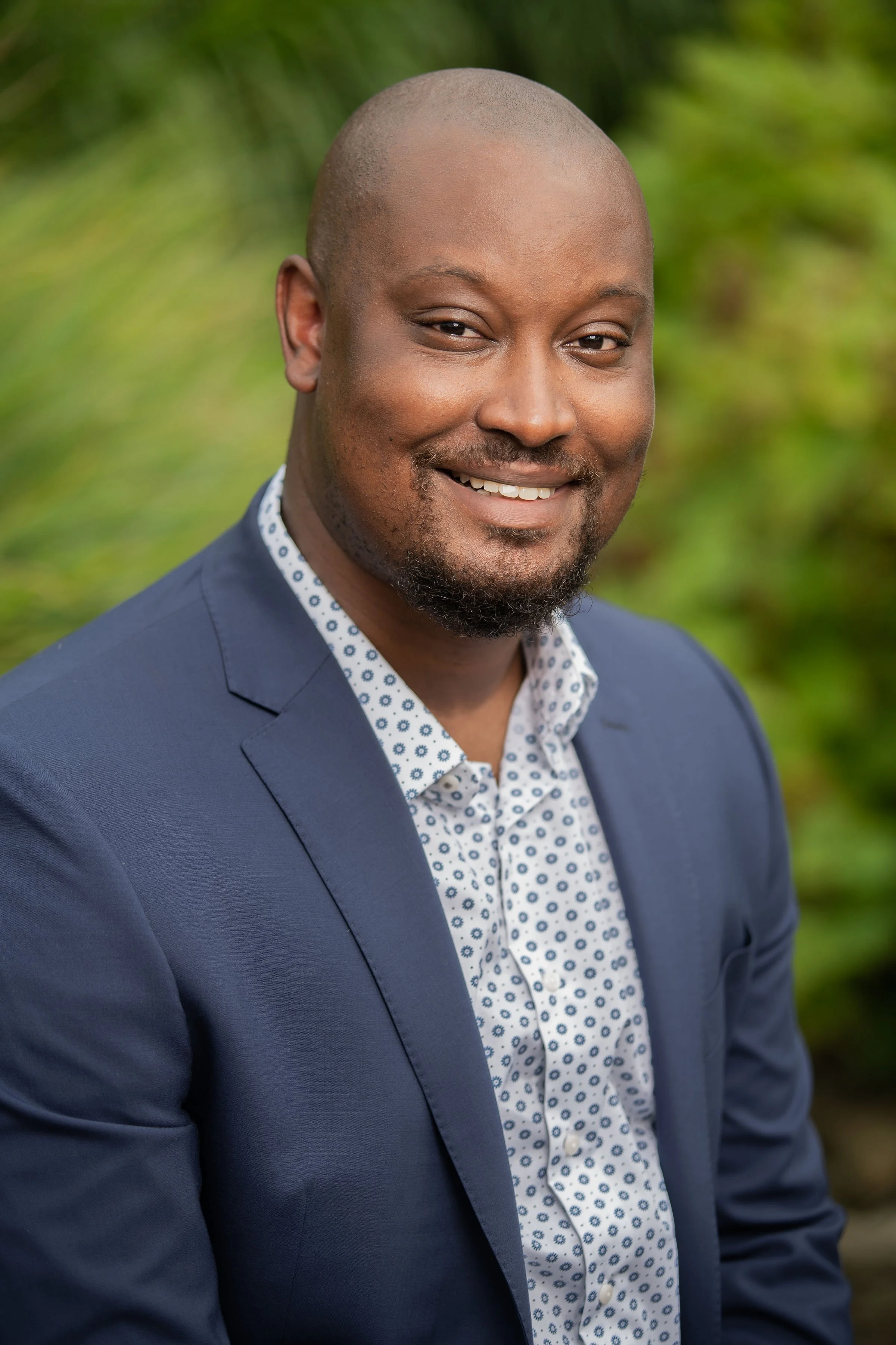 A smiling man with a shaved head and goatee, wearing a blue blazer and a patterned white shirt, standing outdoors with a blurred green natural background.