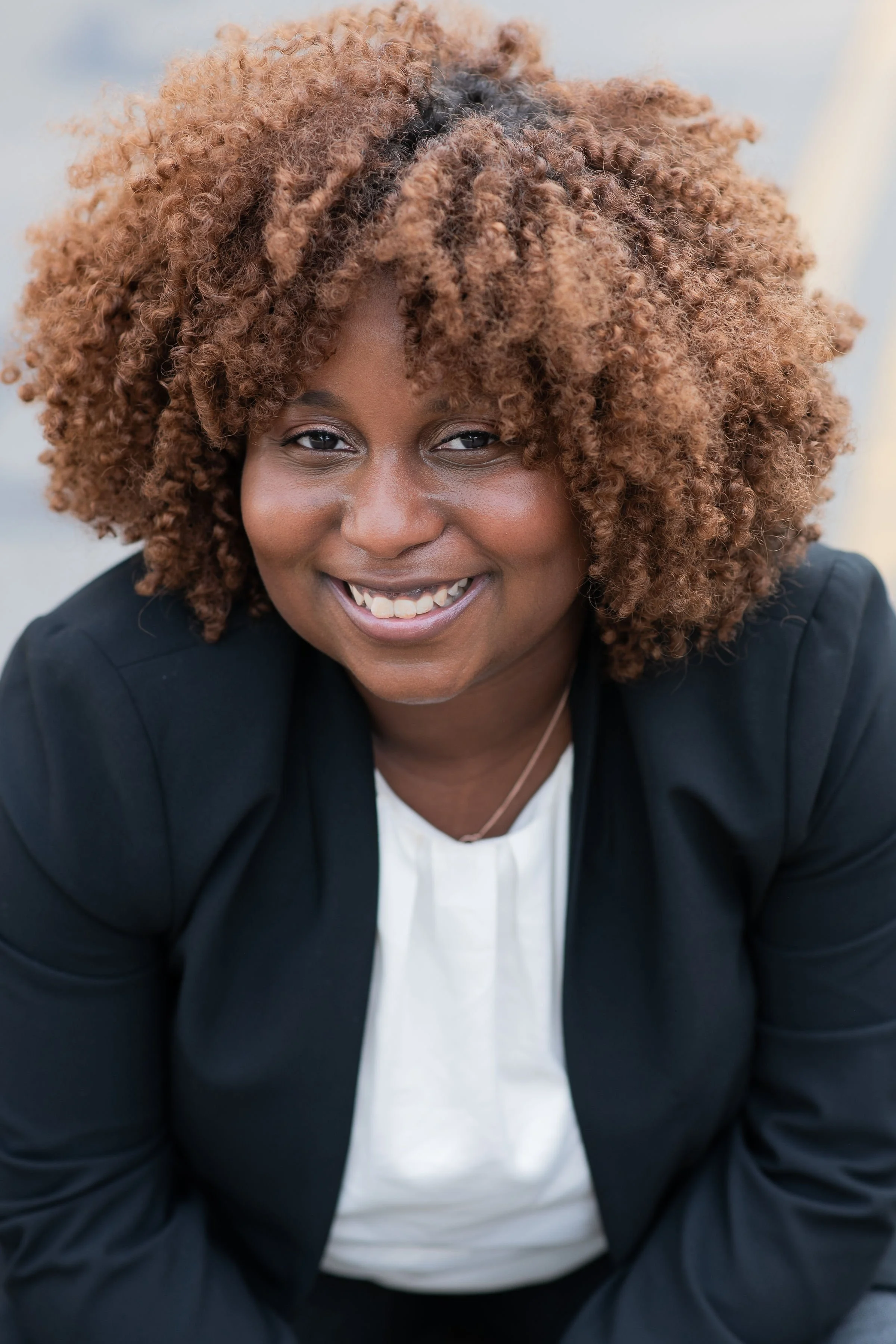 A woman with curly, reddish-brown hair, smiling while looking at the camera, wearing a black blazer over a white shirt.