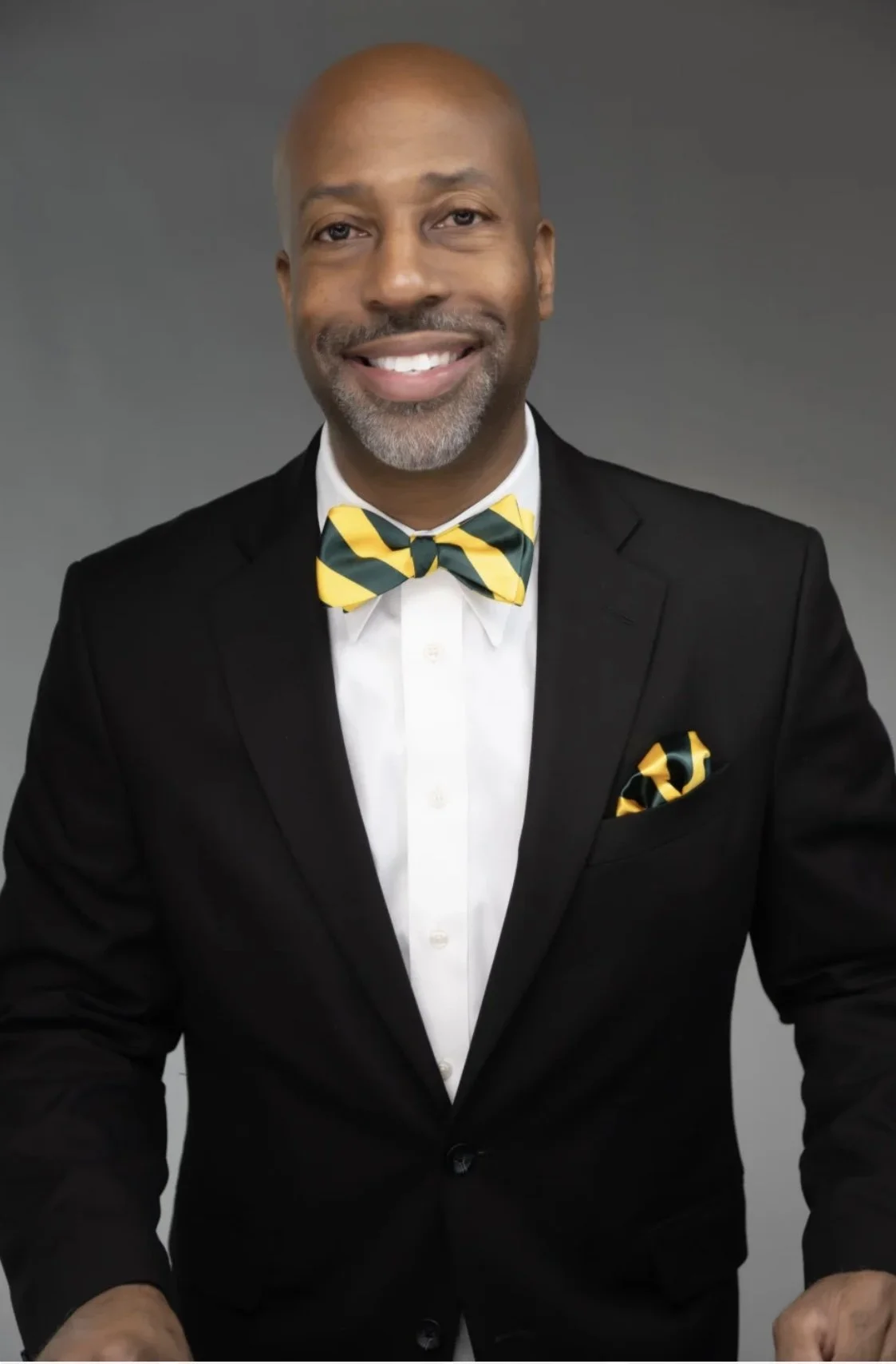 A man in a black tuxedo with a yellow and green striped bowtie and matching pocket square, smiling against a gray background.