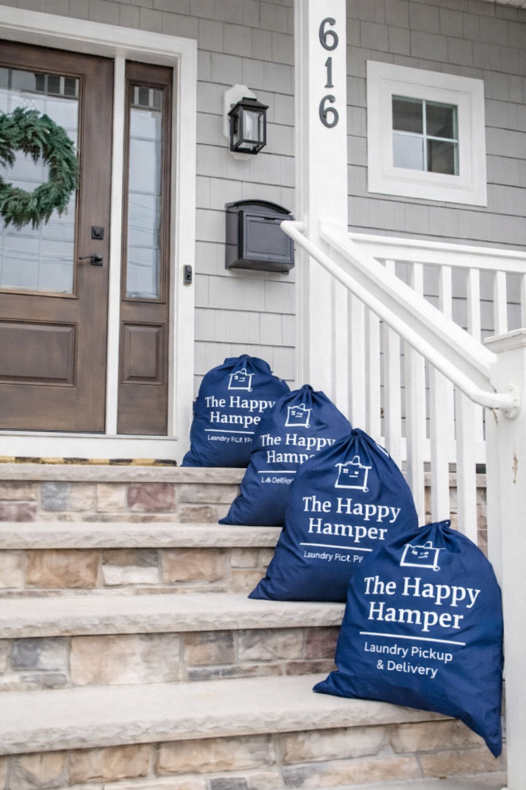 Multiple blue laundry bags labeled 'The Happy Hamper' placed on brick stairs outside a house with a wooden front door, mailbox, and house number 616.