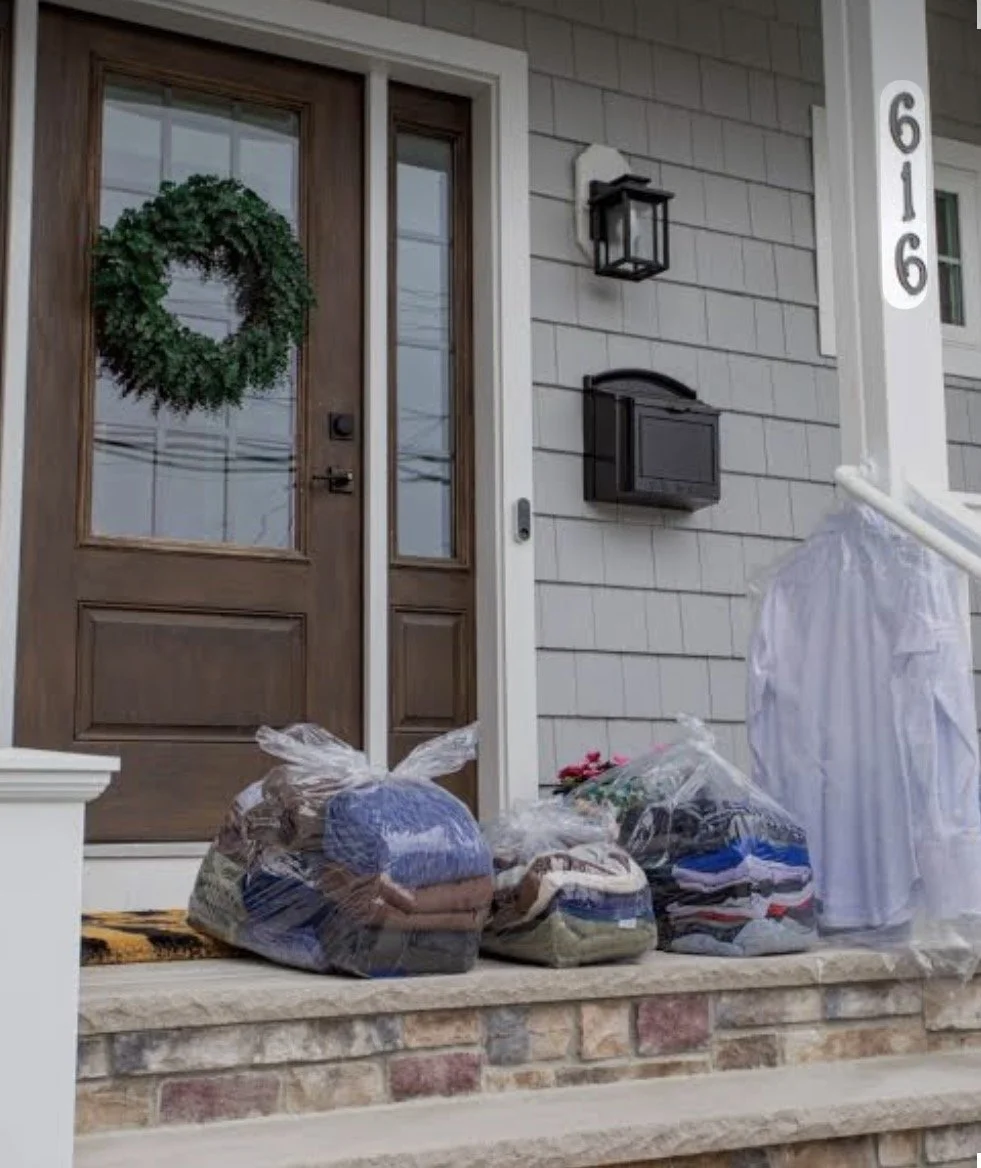 Customers' laundry and personal items in plastic bags placed on the front porch of a house with a brown wooden door and gray siding, with house number 616 displayed on the post.