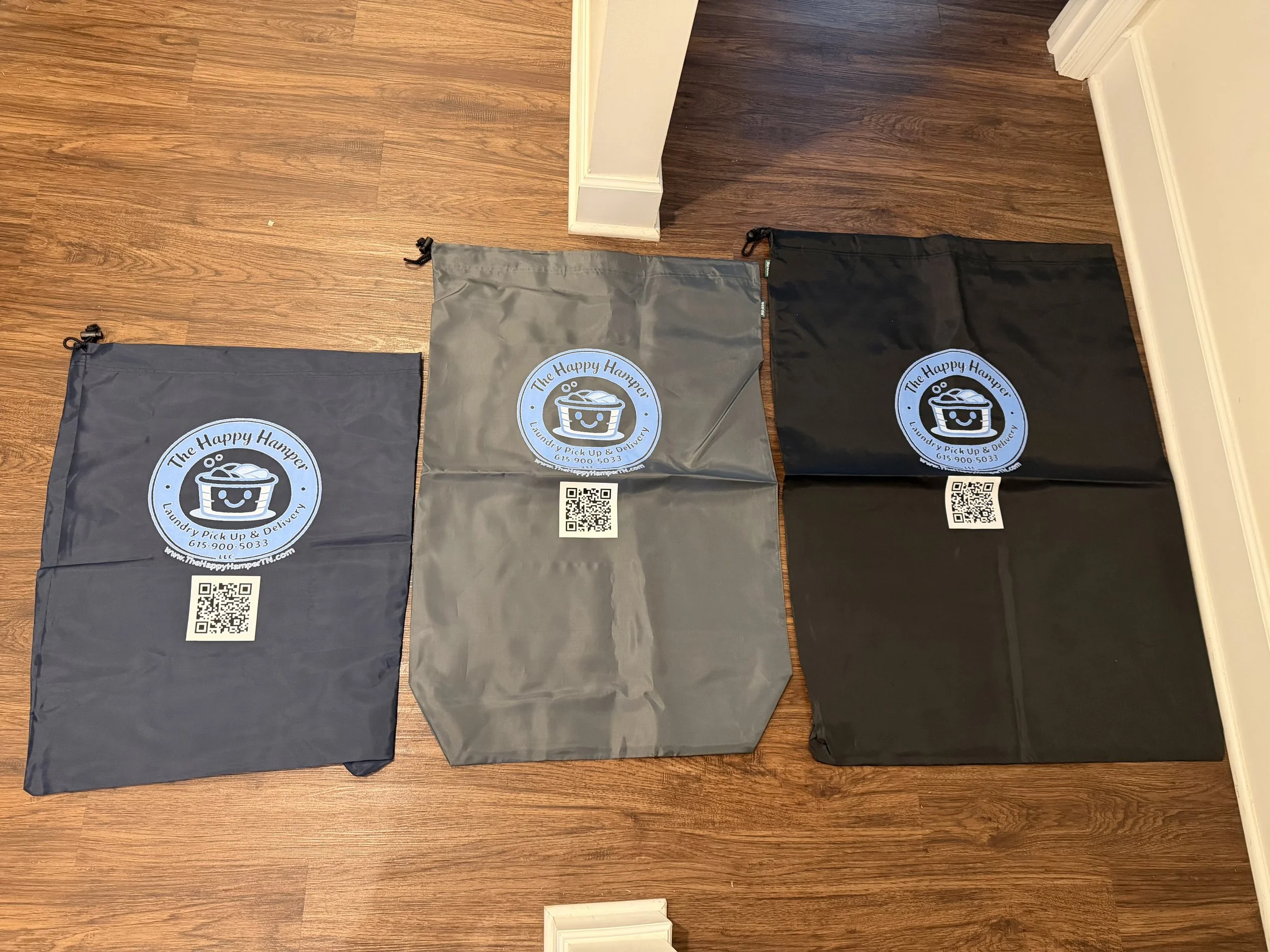 Three laundry bags in black, gray, and dark gray colors with the logo and QR code of 'The Happy Hampers.'