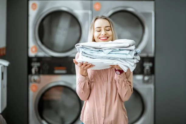 A woman smiling and holding a large pile of folded laundry in front of commercial dryers in a laundry facility.