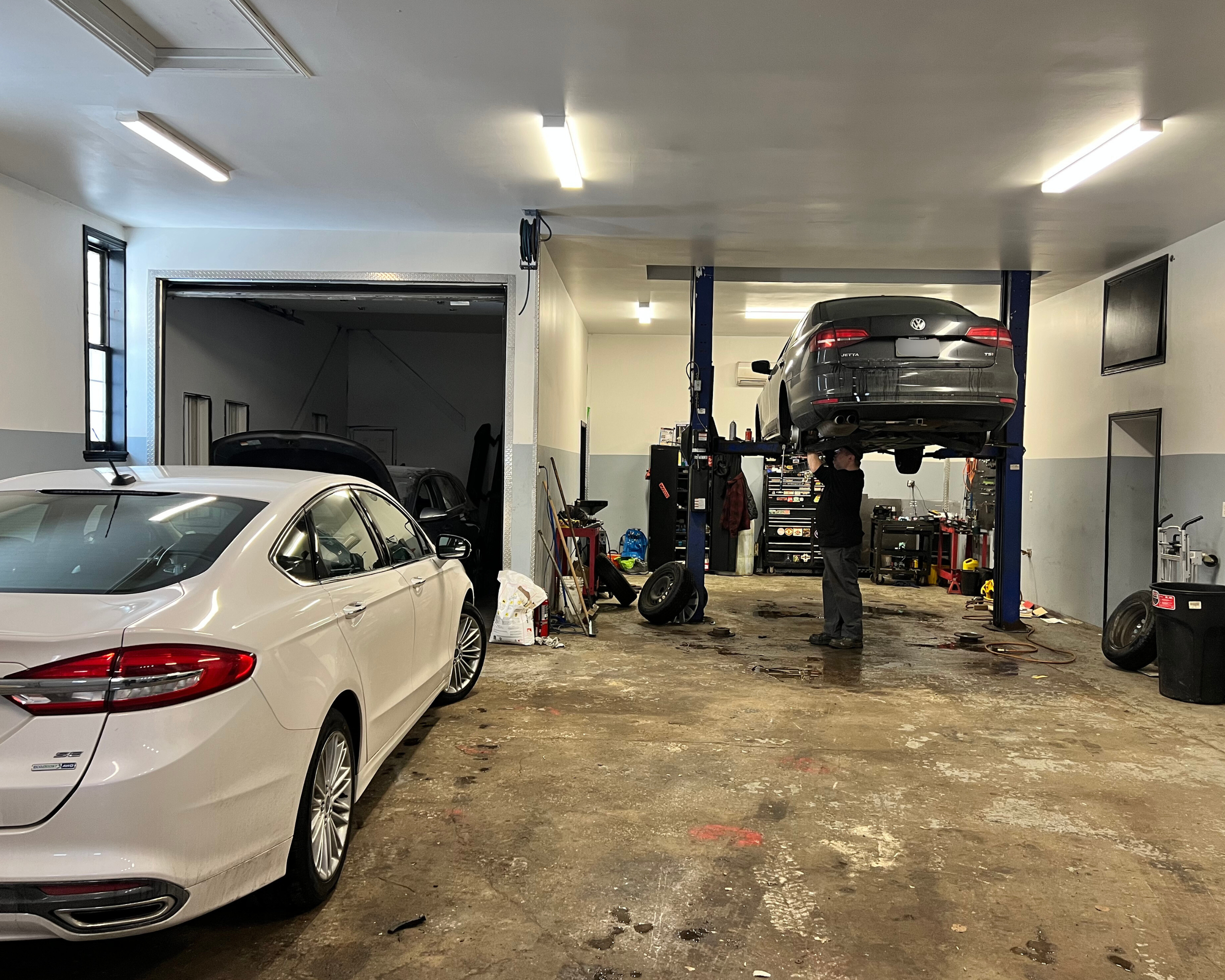 Auto repair garage with a white car on the left and a black car elevated on a lift being worked on by a mechanic in black. Tools and equipment are around the garage.