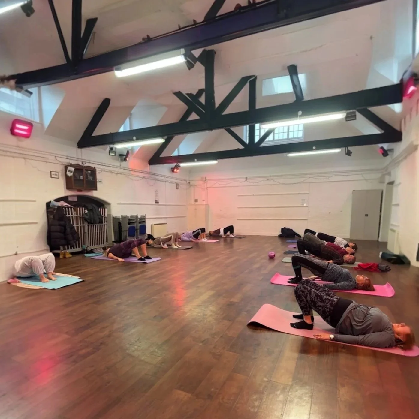 A group of people participating in a yoga class, lying on yoga mats in a spacious, well-lit studio with wooden floors and exposed ceiling beams.