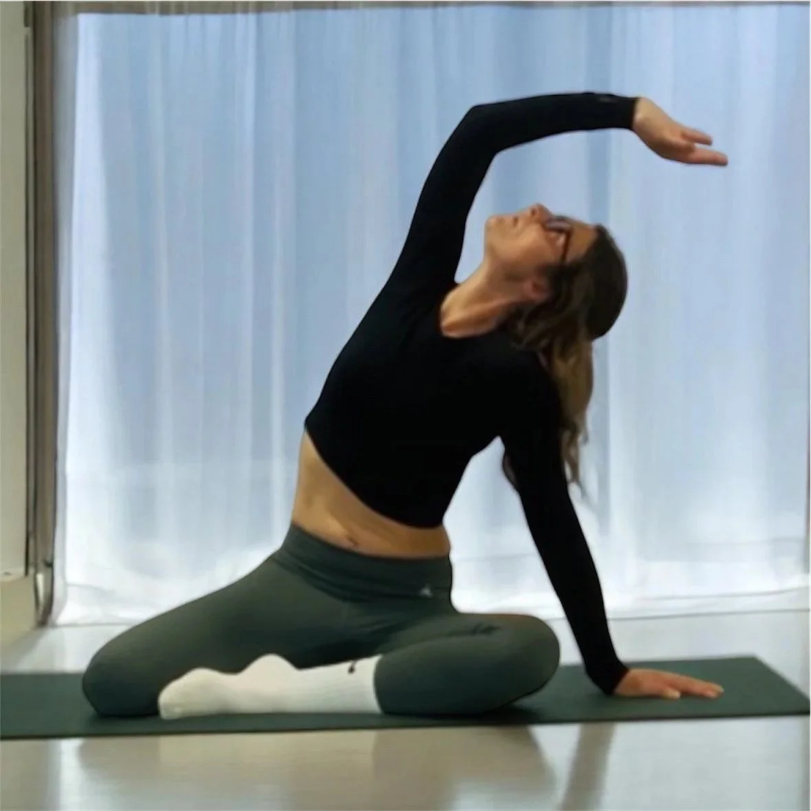 Woman in black long-sleeve shirt and gray leggings practicing yoga indoors, sitting on a mat in a spinal twist pose with a white curtain in the background.