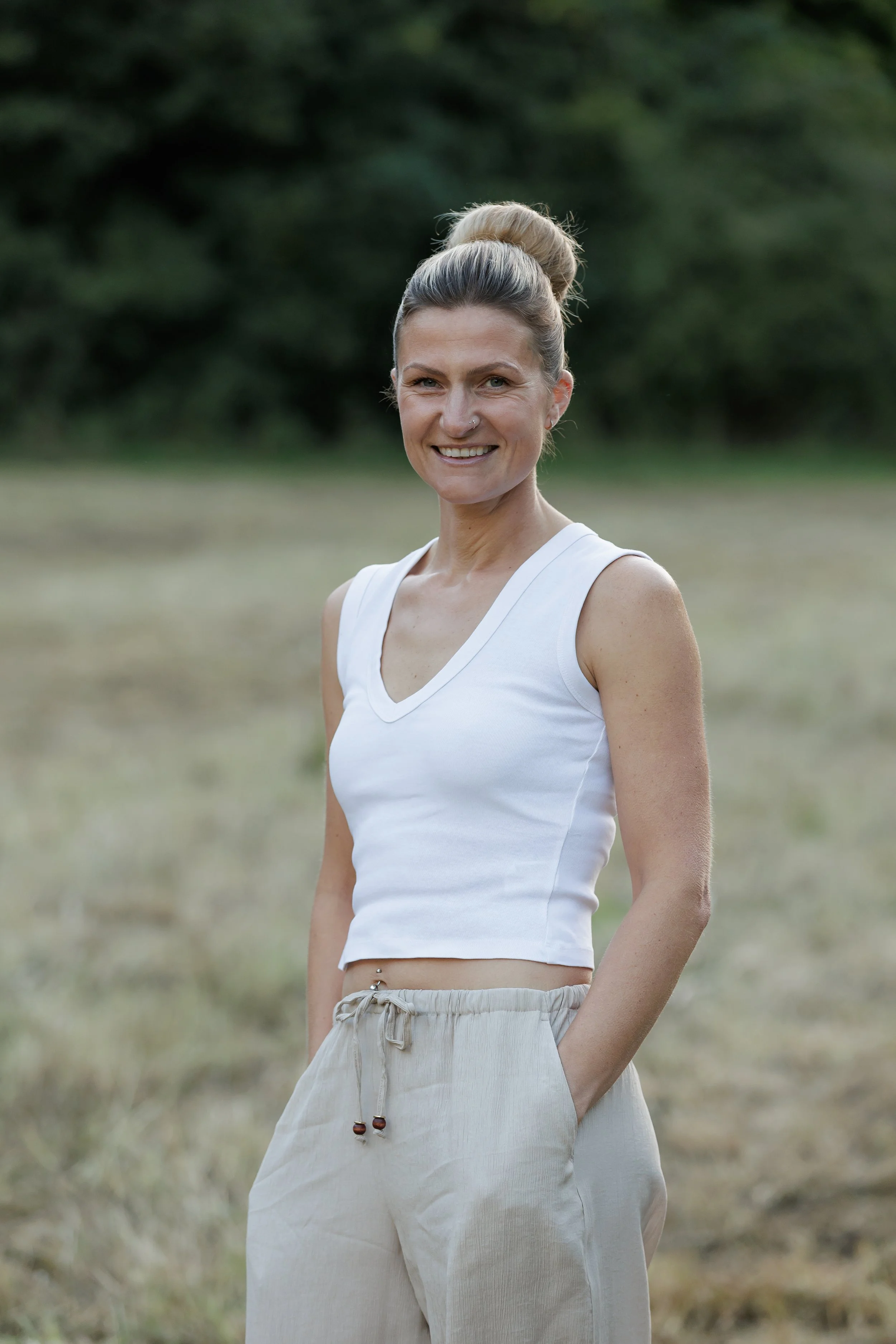 A woman standing outdoors in a field, smiling at the camera, with her hair in a bun and wearing a sleeveless white top and light-colored pants.