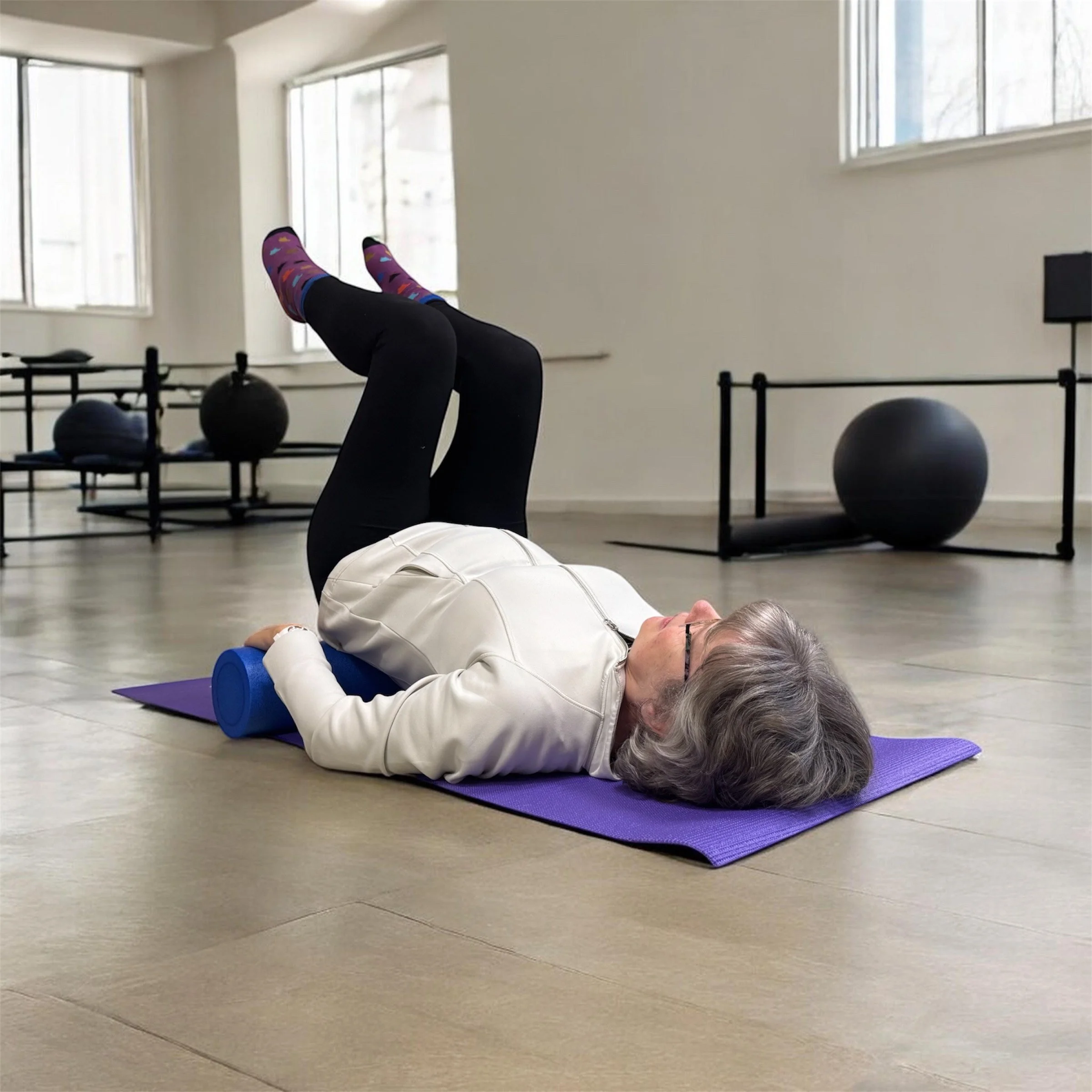 Elderly woman lying on her back on a purple exercise mat with a foam roller under her neck, practicing stretching or yoga in a bright fitness studio with large windows and exercise equipment in the background.