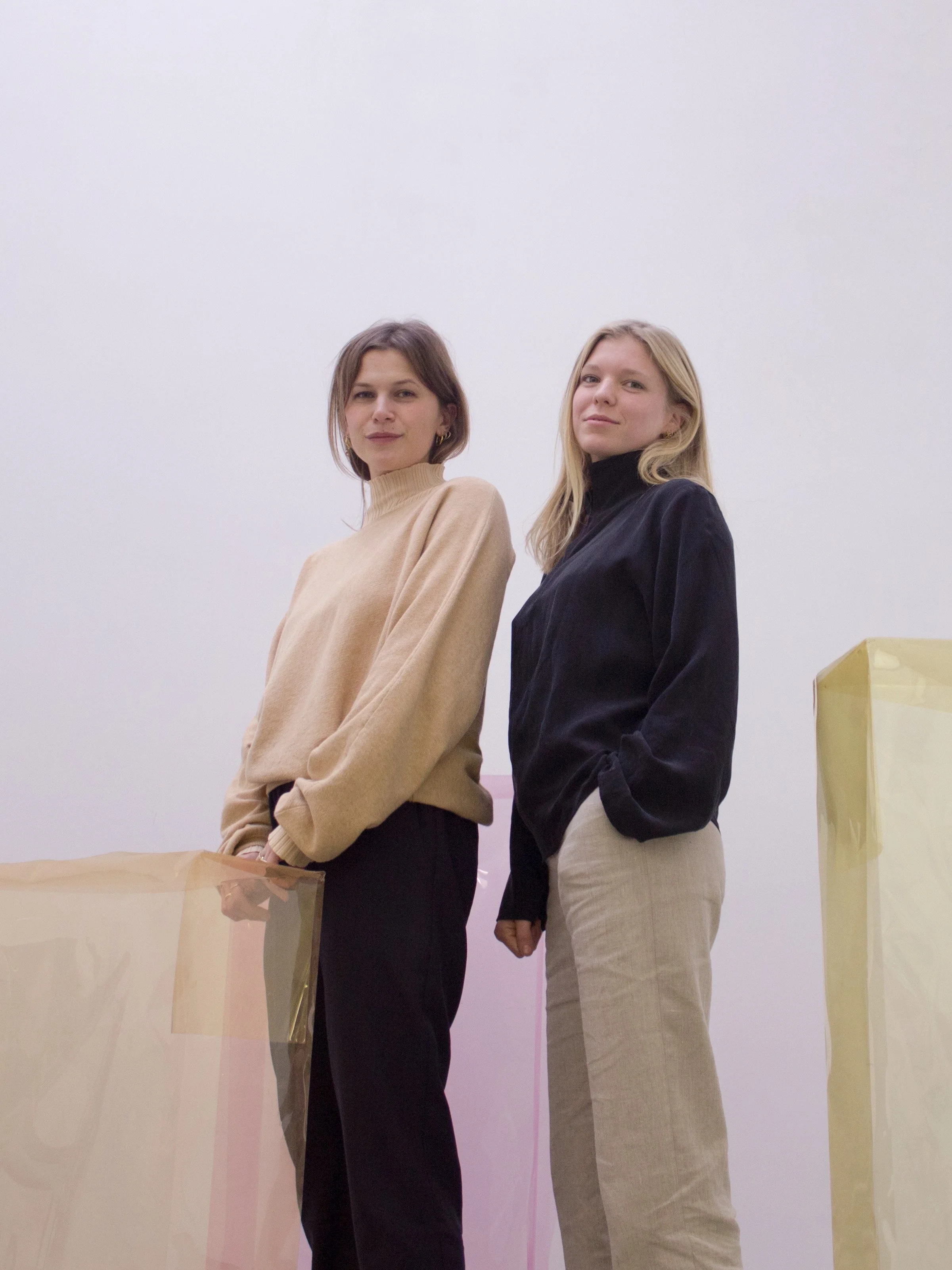 Two women standing in front of a white background, wearing sweaters and pants, with transparent colored sheets around them.