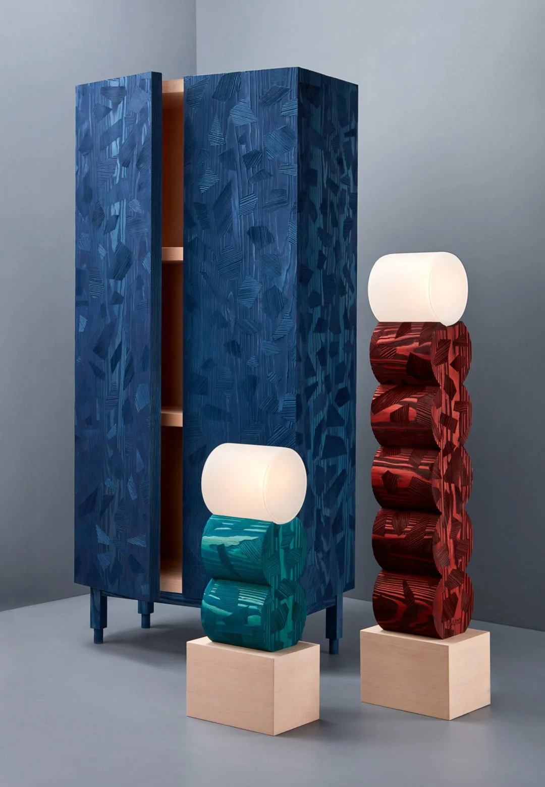 A textured blue cabinet with open door beside two modern sculptural lamps. The lamps feature cylindrical shapes, with one lamp having turquoise cylinders and the other red, each topped with a frosted bulb, set on wooden bases.