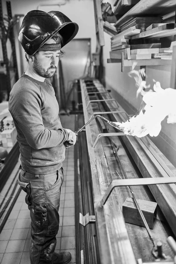 Black and white image of a man in a workshop using a torch, with a welding helmet raised on his head, standing by a workbench with wooden planks and metal bars.
