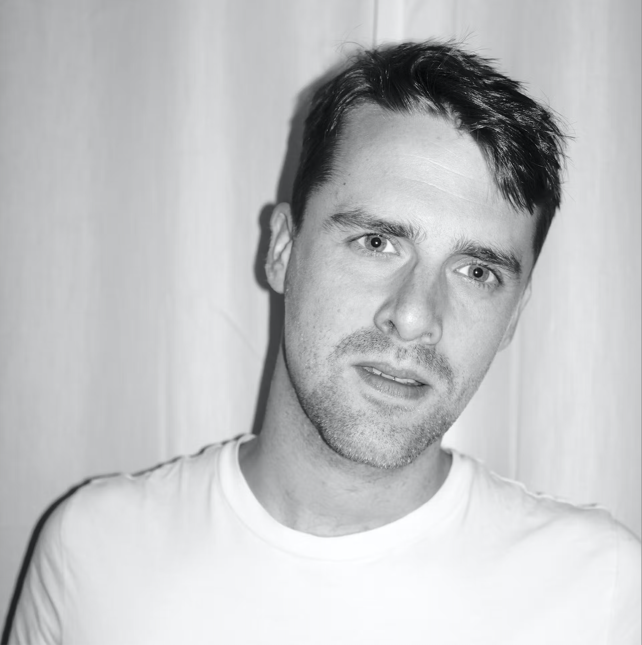 Black and white portrait of a man wearing a white t-shirt, with short hair and a neutral expression.