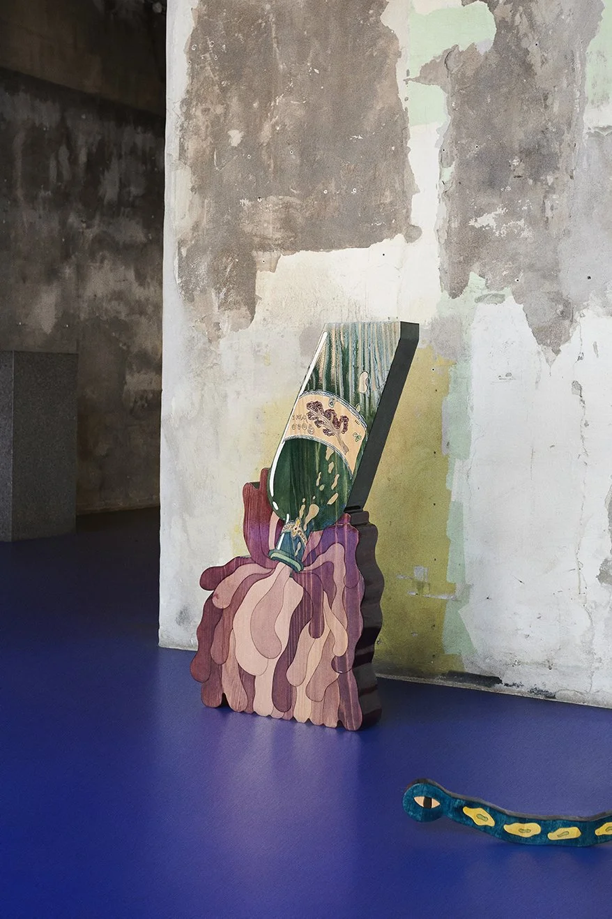 Artistic wooden sculpture of a champagne bottle pouring liquid on a blue floor, placed against a weathered wall.