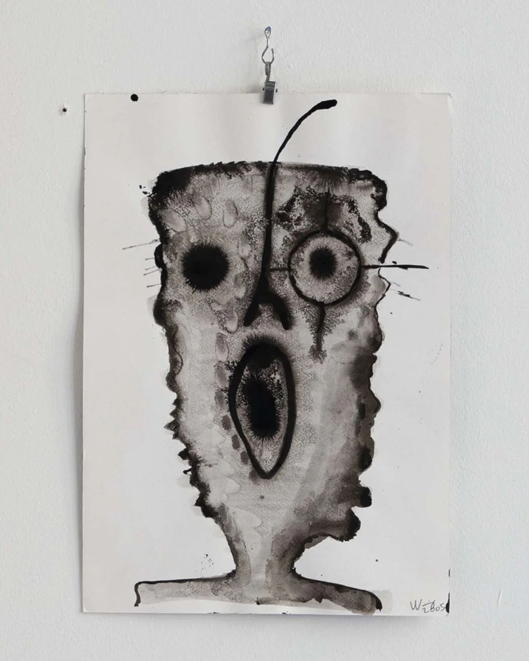 Abstract ink drawing resembling a face with large eyes and an open mouth, mounted on a wall with a clip.