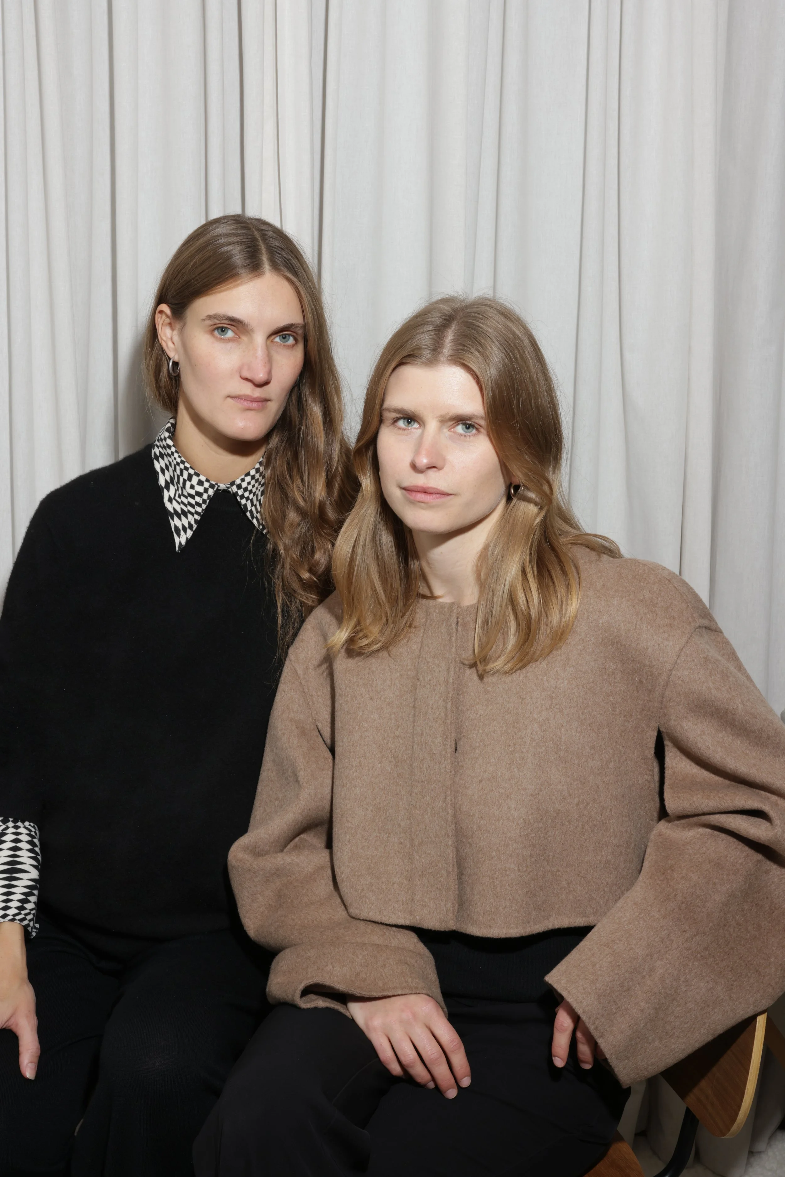 Two women sitting in front of white curtains, one wearing a black outfit with a patterned collar, and the other in a beige jacket.