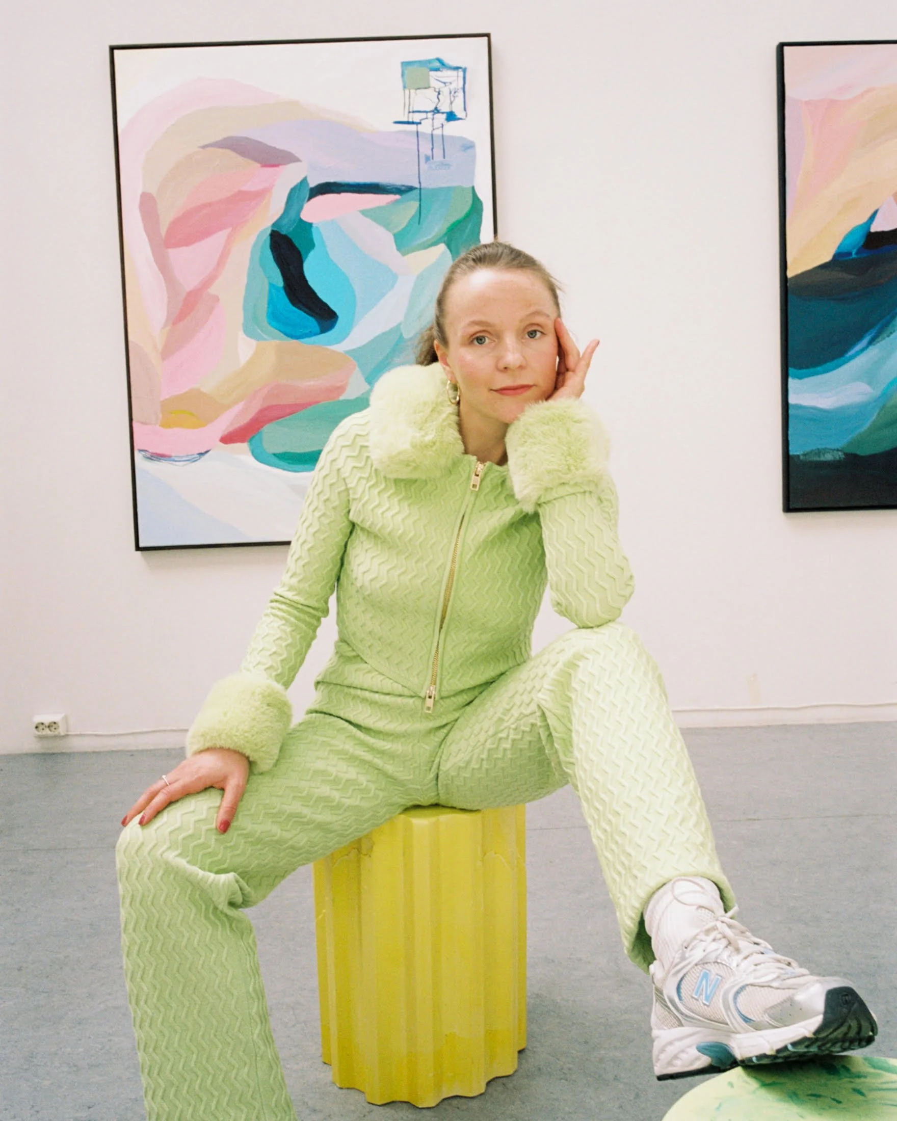 Person in a green textured outfit and sneakers sitting on a yellow stool in an art gallery, surrounded by abstract paintings on the walls.