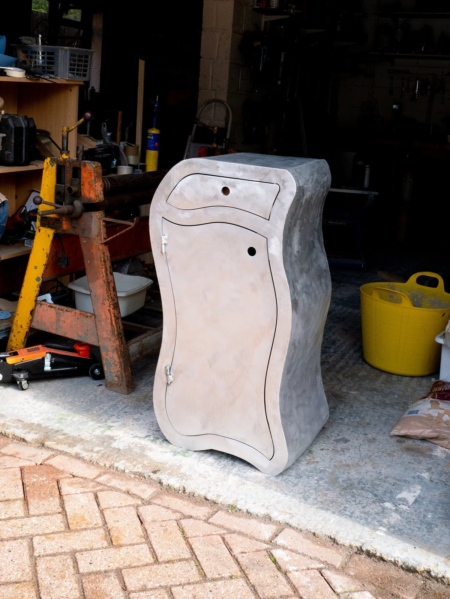 A wooden cabinet with a distorted, wavy design placed outside a workshop with tools and equipment in the background.