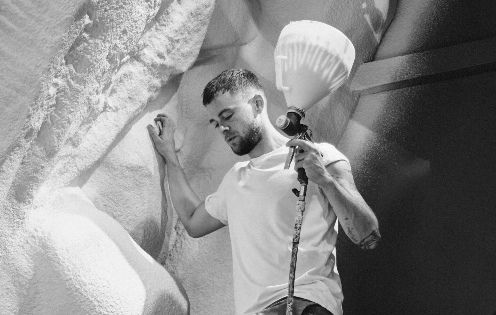 Man in a white t-shirt holding a spray paint gun, standing next to a textured wall.