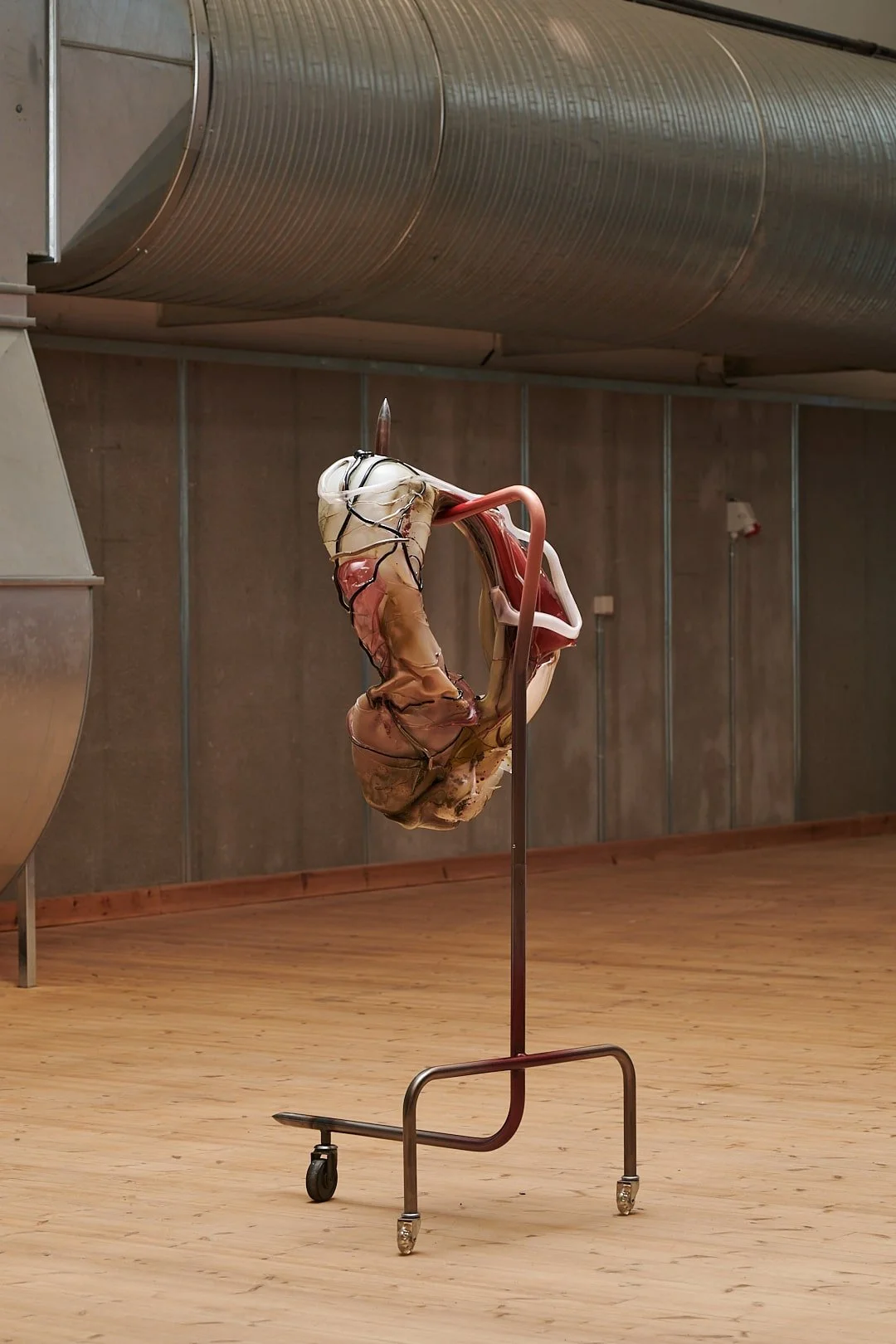 Art installation featuring a distorted, organic sculpture suspended on a metal stand with wheels, displayed in an industrial setting.