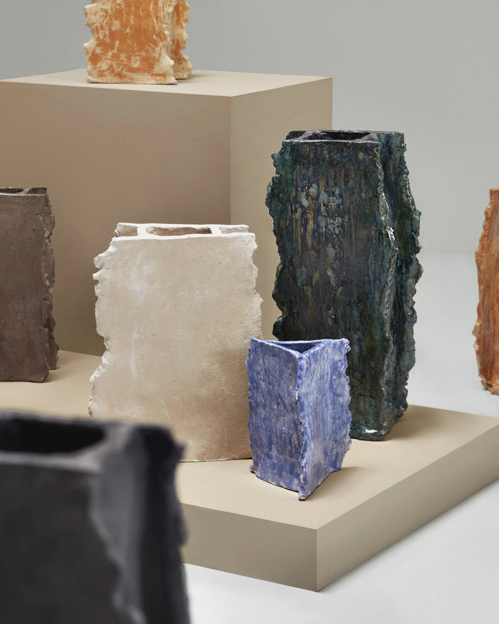 Abstract ceramic vases on display, featuring rough textures and various colors including white, blue, orange, green, and black, arranged on a beige pedestal.