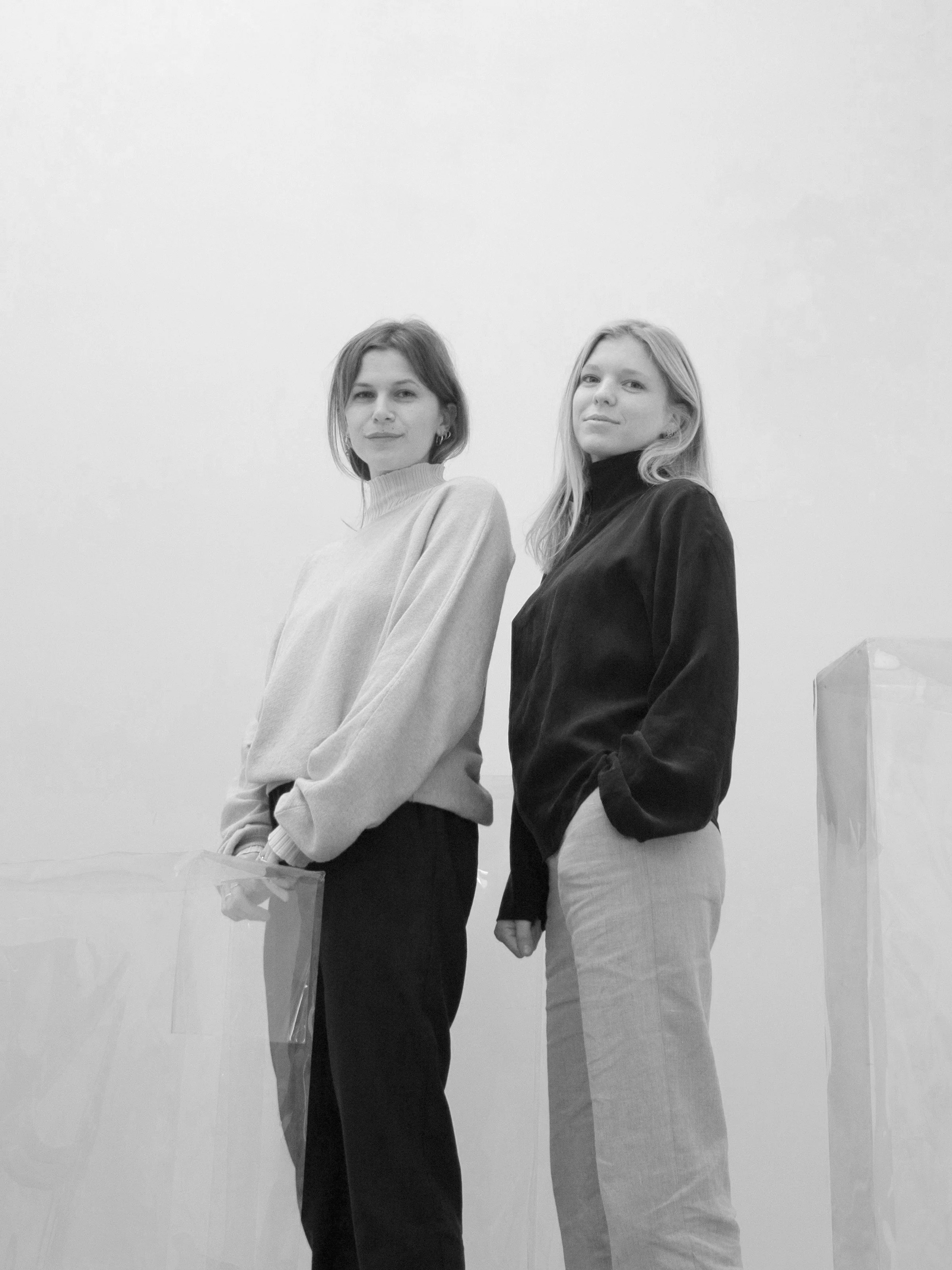 Two women standing in a minimalist setting, both wearing sweaters and pants, looking at the camera in a monochrome photograph.