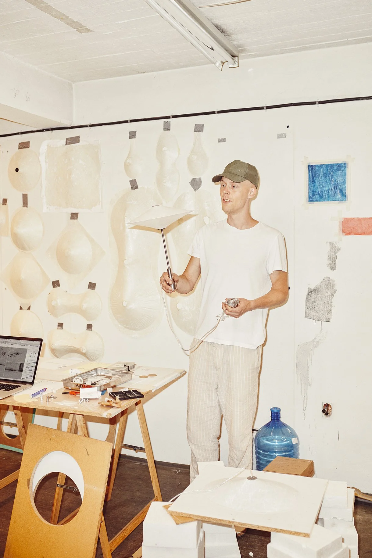 Person in a workshop holding a tool, surrounded by abstract wall art and a cluttered table with a laptop and tools.