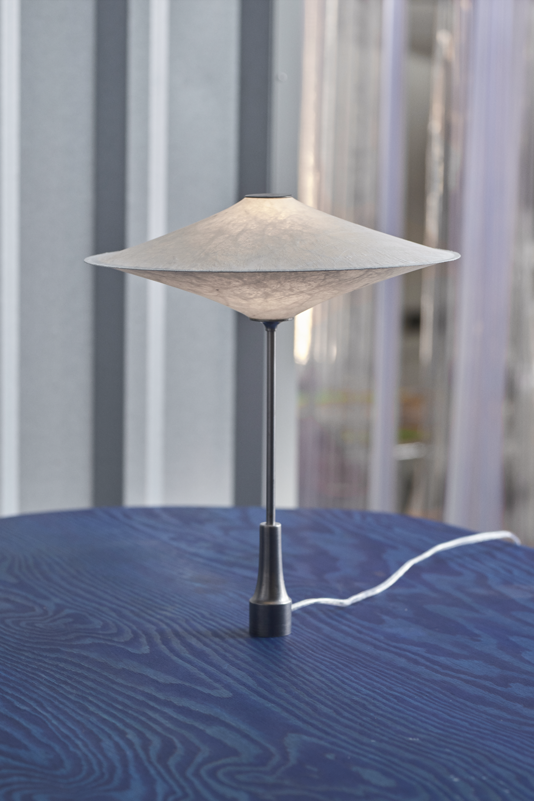 A modern table lamp with a wide, conical paper shade and a slender metal base, positioned on a blue wooden surface.