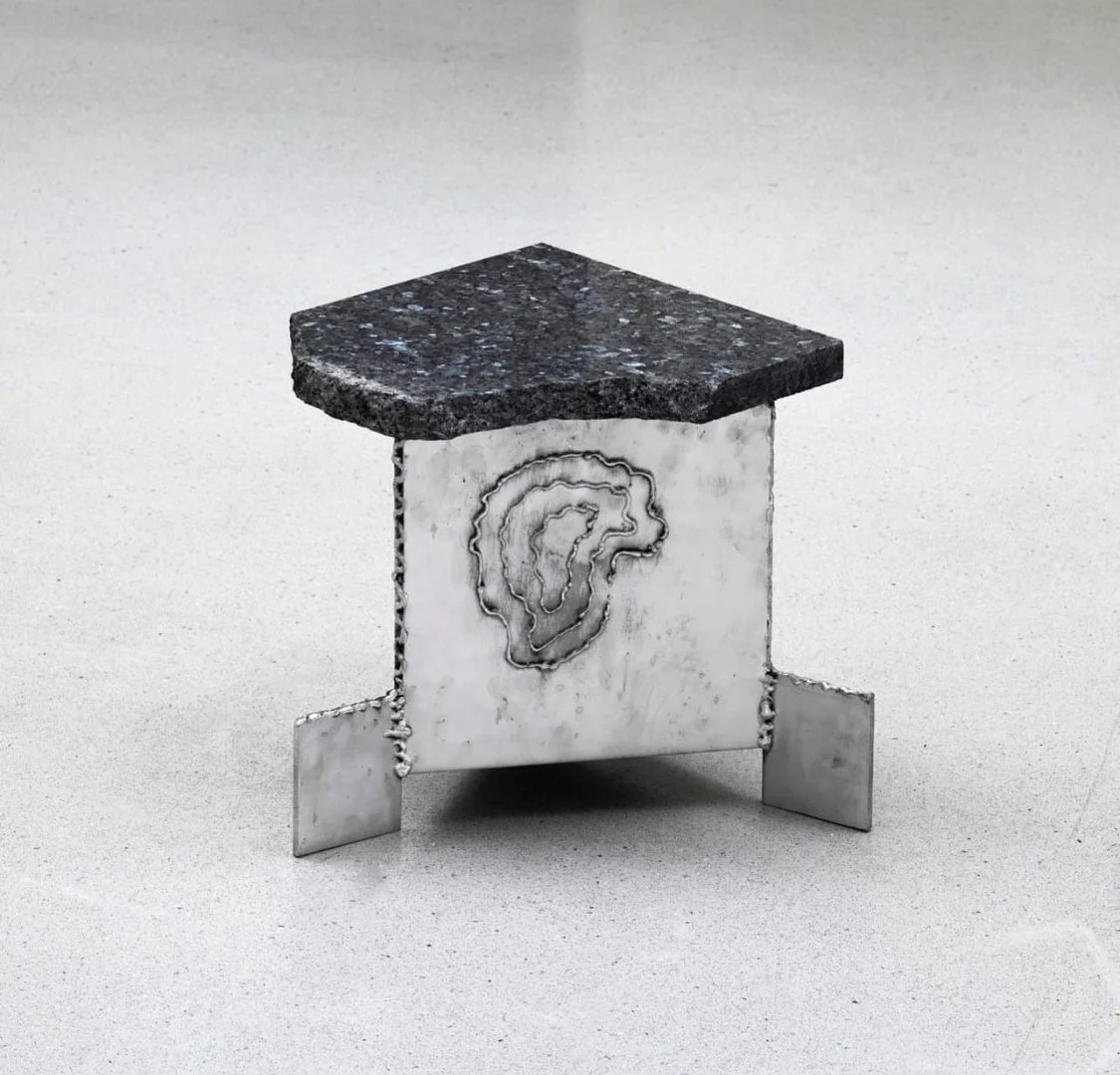 A geometric stool with a textured metallic base and a dark stone top, placed on a white surface.