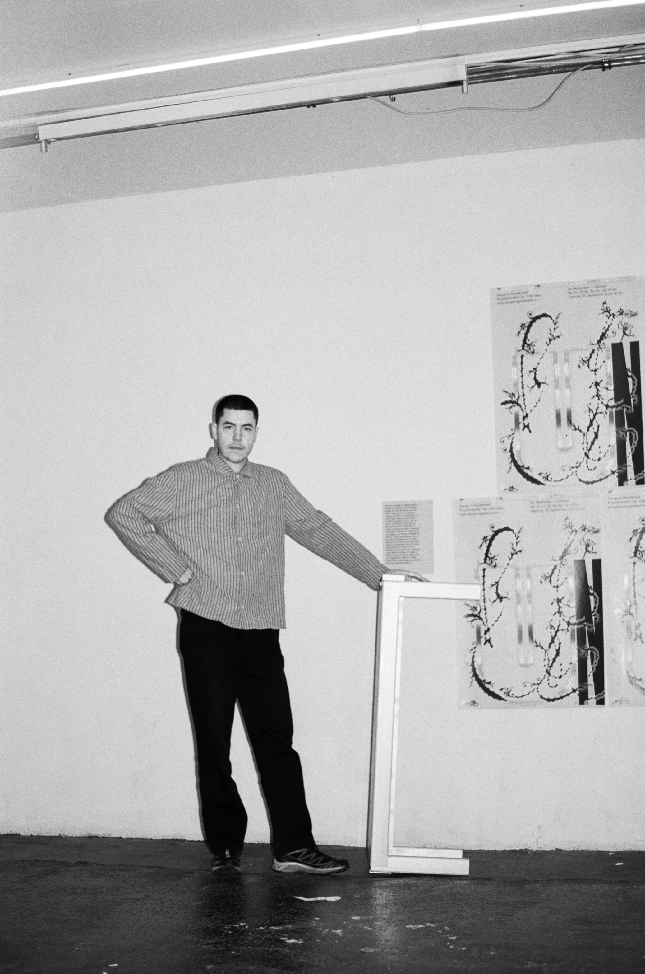 Black and white photo of a person standing indoors next to abstract wall art. They are wearing a striped shirt and dark pants, positioned with one hand on their hip and the other resting on a frame. The background features a plain wall with artwork attached to it.