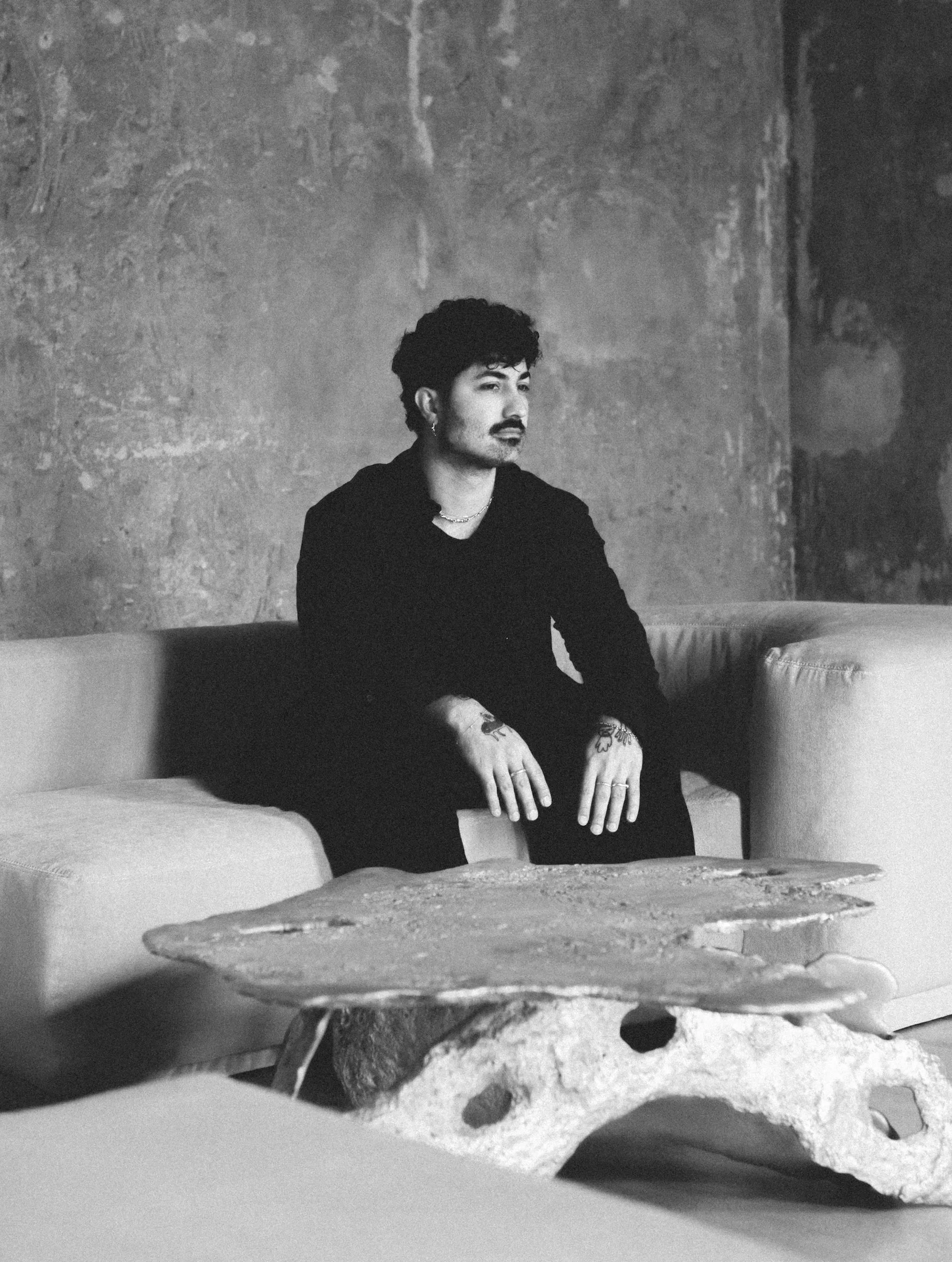 Black and white photo of a man sitting on a couch with a textured stone coffee table in front, against a rough wall.
