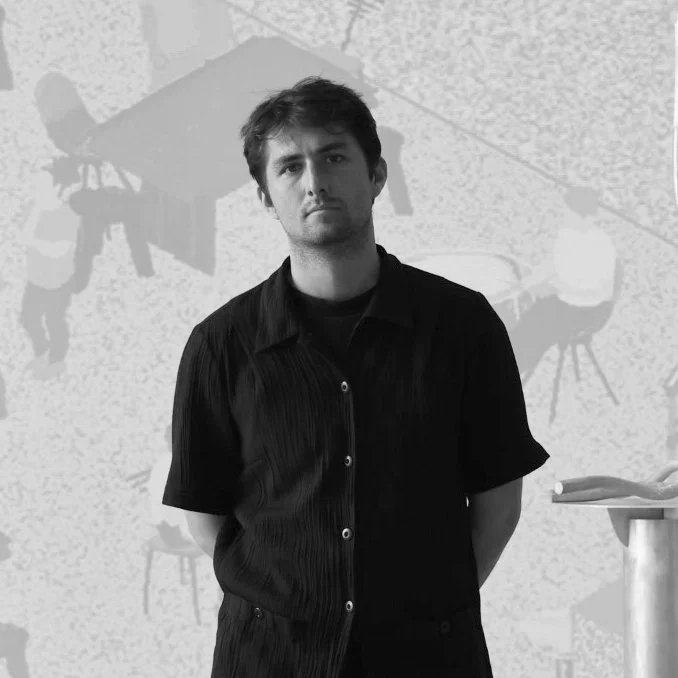 Black and white photo of a person standing indoors, wearing a dark shirt. The background shows a wall with an abstract design or mural depicting chairs and tables.