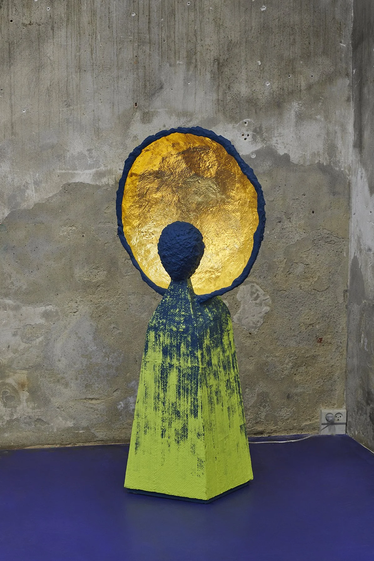 A yellow and blue modern sculpture with a textured, abstract form and a circular gold disk behind it, set against a concrete wall.