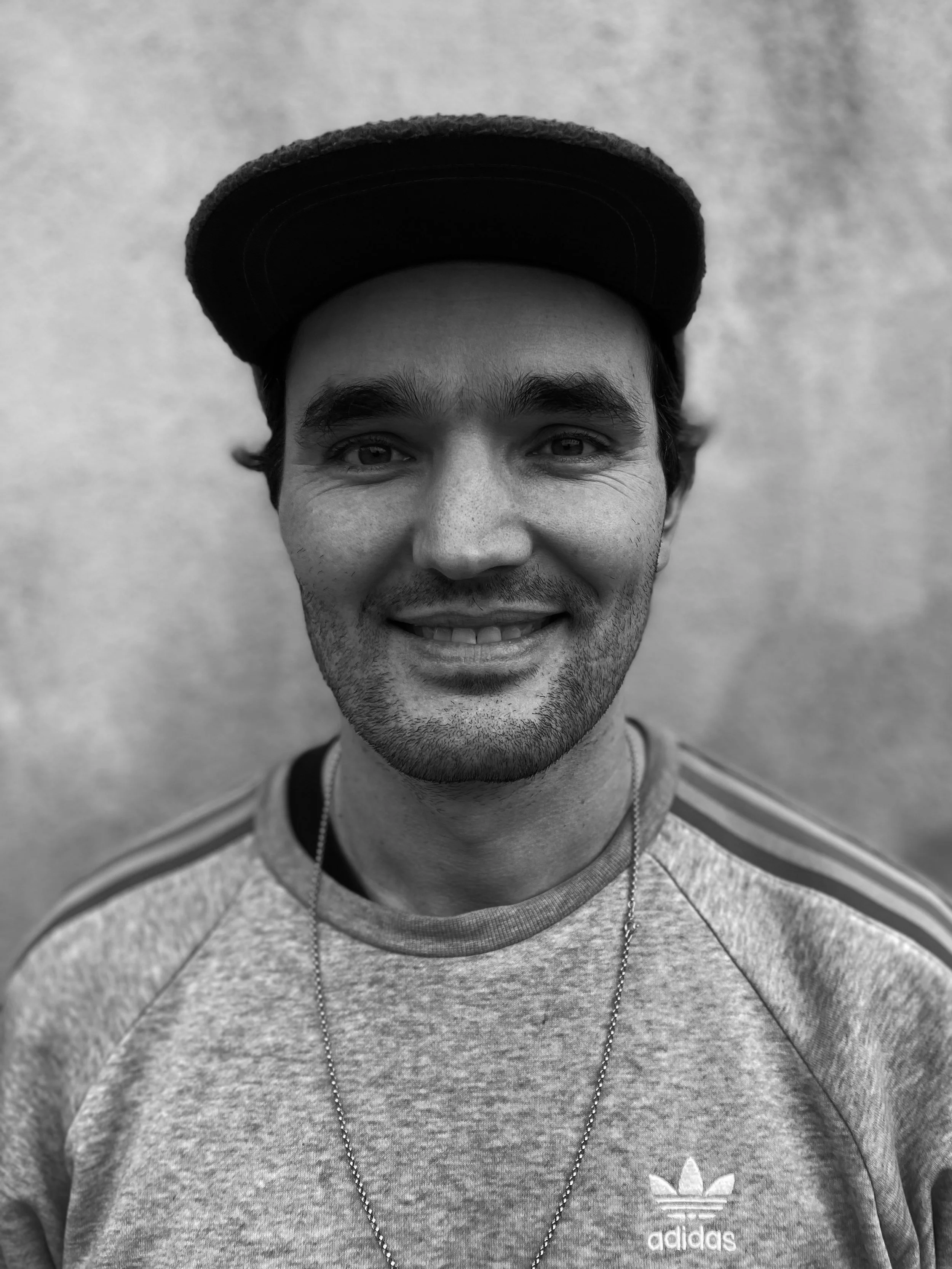 Black and white photo of a person wearing a hat and an Adidas sweatshirt, smiling at the camera.