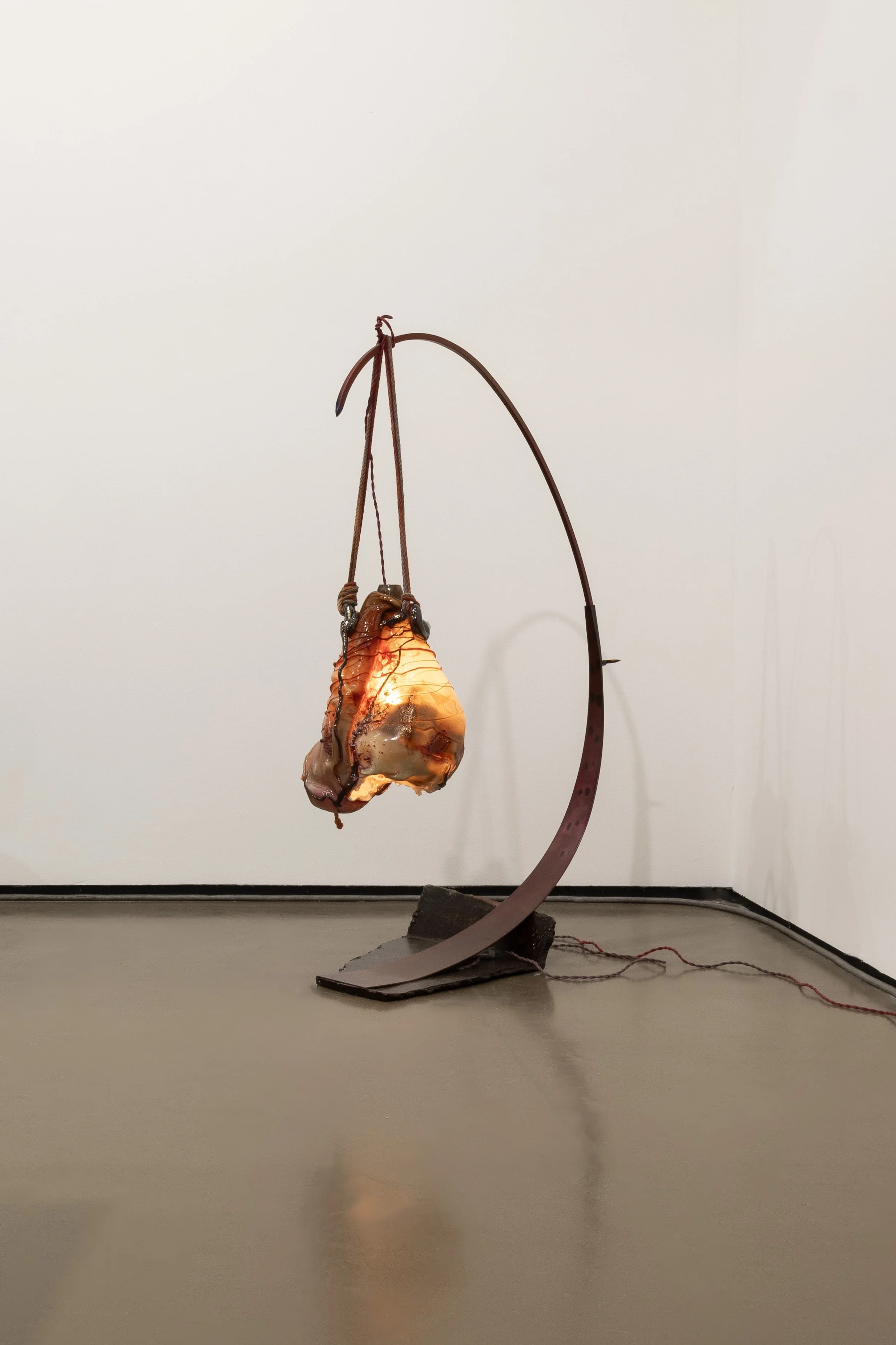 Abstract sculpture with a suspended, illuminated organic shape on a curved metal stand in a gallery setting.