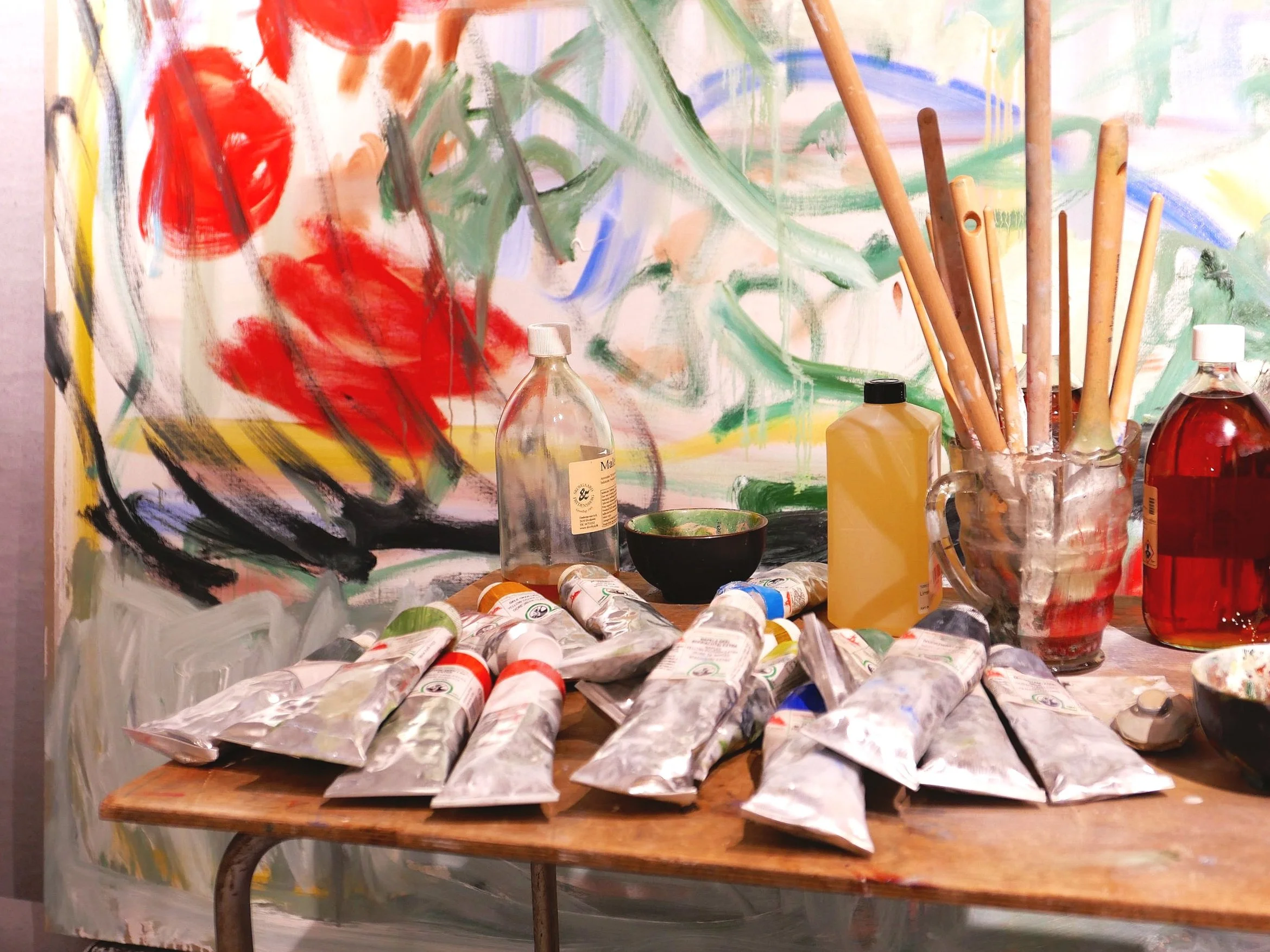 Assorted artist paint tubes, brushes in a jar, and bottles on a wooden table with a colorful abstract painting in the background.