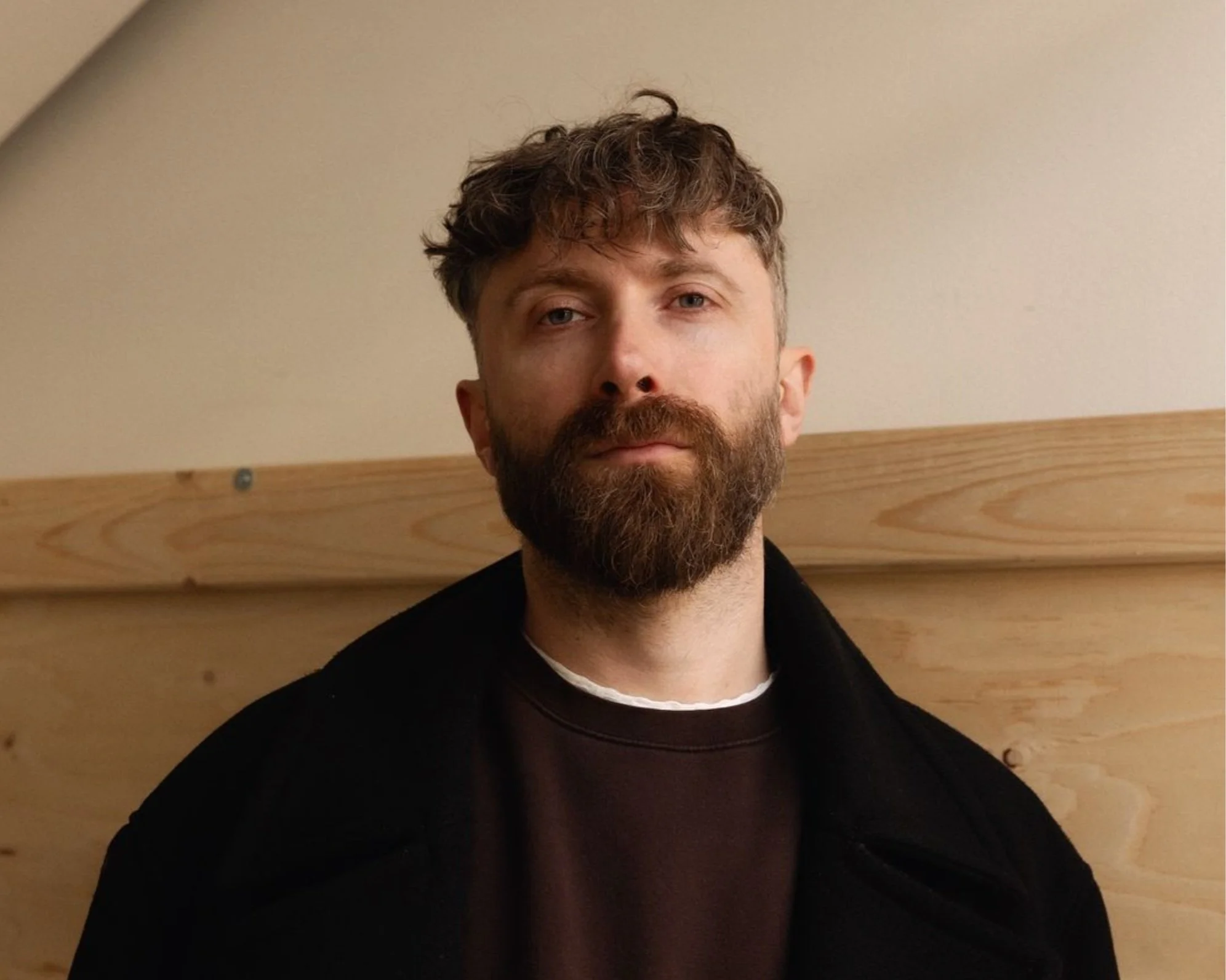 A bearded man with short curly hair wearing a black coat and dark shirt, standing in front of a wooden background.