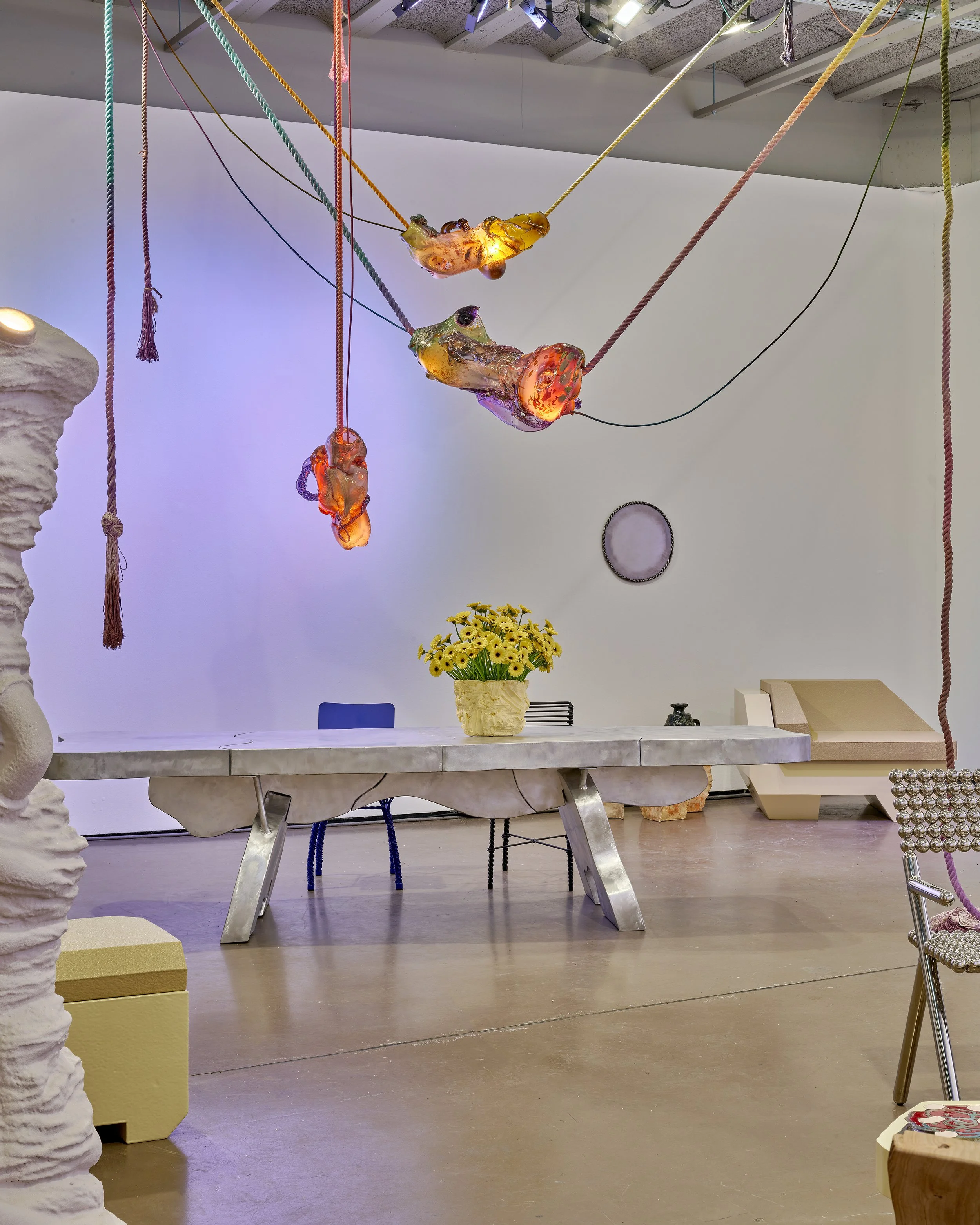 Modern art installation with abstract sculptures, a metal table with a yellow flower centerpiece, colorful chairs, and hanging lights from ropes against a white wall.