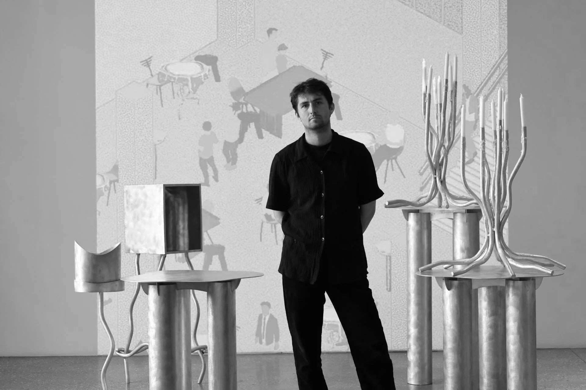 Black and white photo of an art gallery with a person standing among abstract metal sculptures on pedestals and a mural in the background.