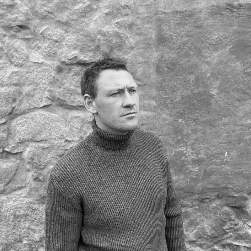Black and white photo of a person in a turtleneck sweater standing in front of a stone wall.