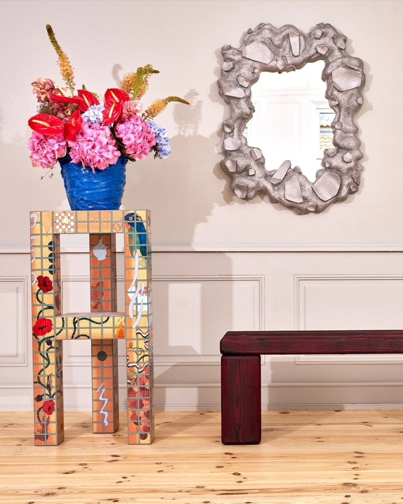Artistic interior featuring a textured mirror and a mosaic-patterned pillar with a blue vase containing vibrant flowers, next to a deep red wooden bench on a wooden floor.